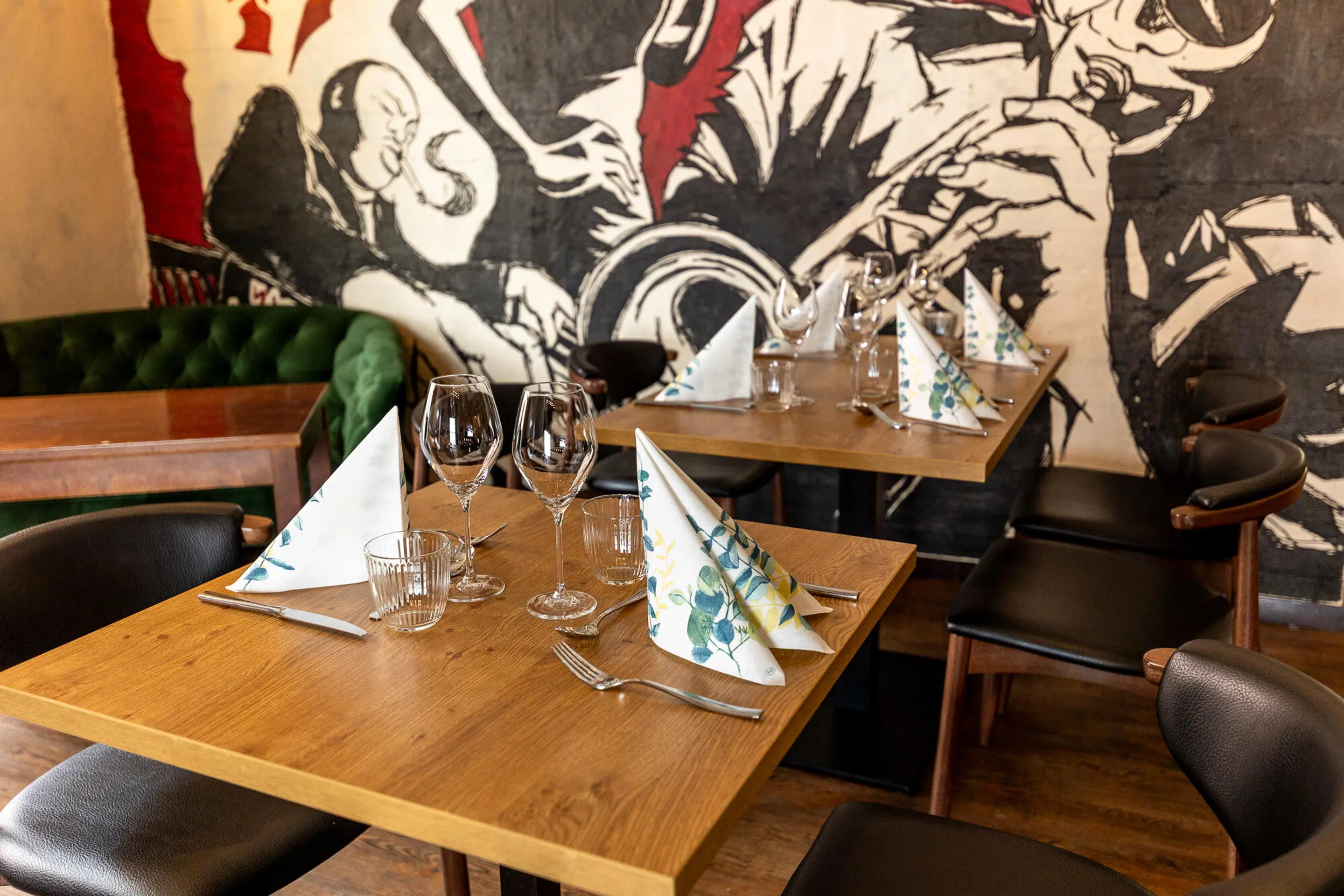 Restaurant tables set with wine glasses and napkins. In the background, a black and white jazz-themed wall mural and a green velvet bench.