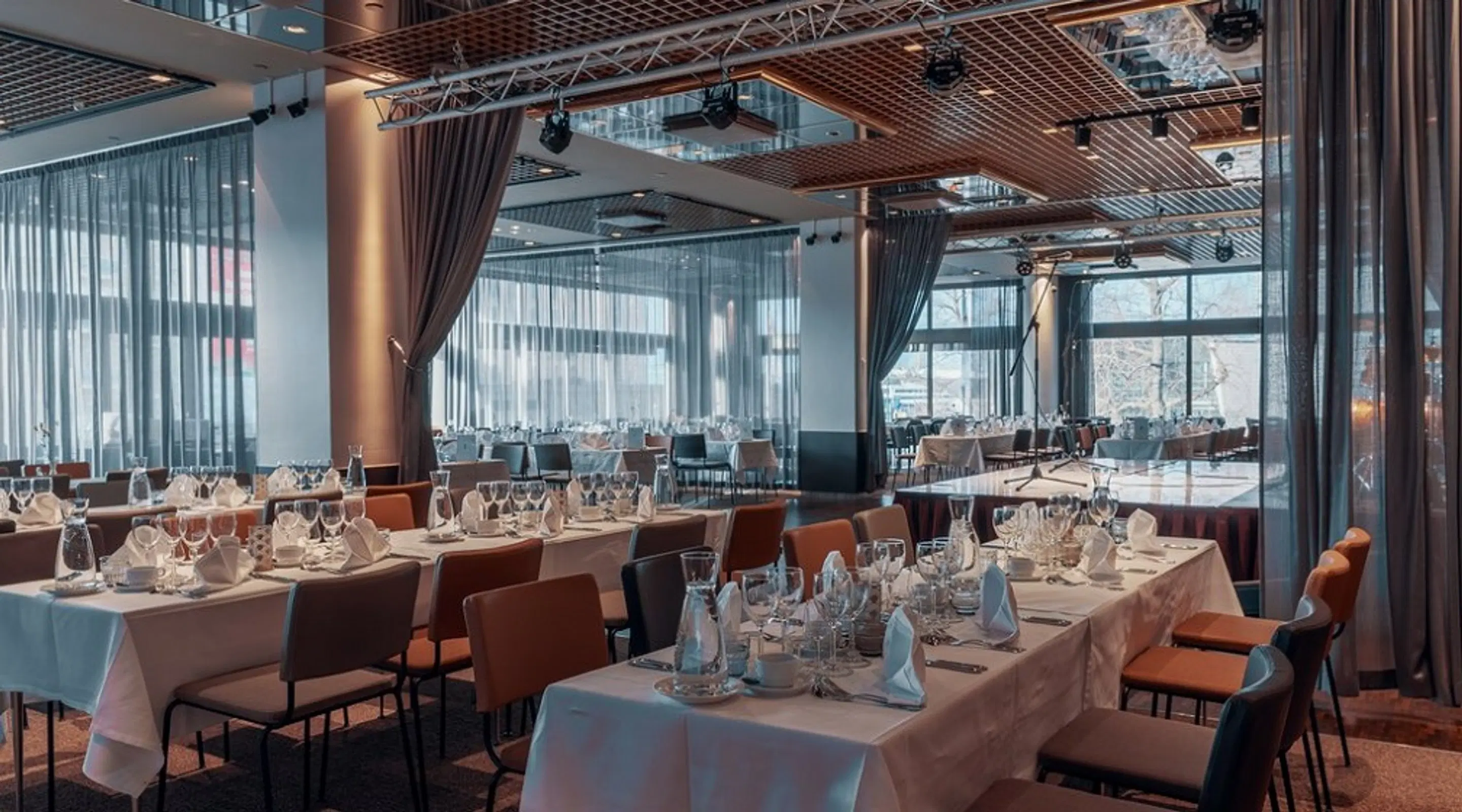 Modern restaurant interior with white tablecloths and orange chairs. Glass walls, gray curtains, and wooden ceiling elements throughout the space.