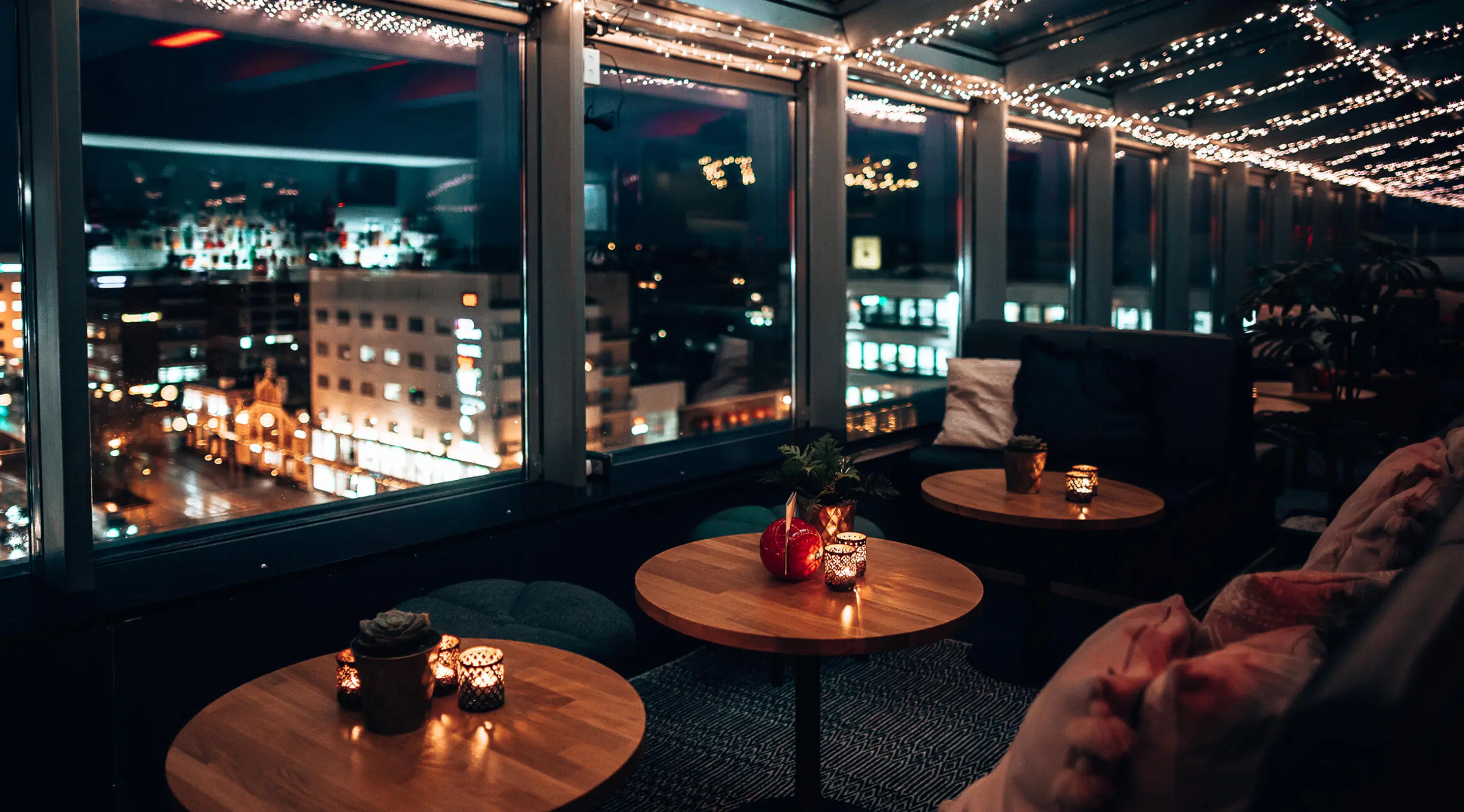Atmospheric rooftop lounge with wooden tables, sofas and candlelight. City lights visible through windows at night, decorative string lights on ceiling.