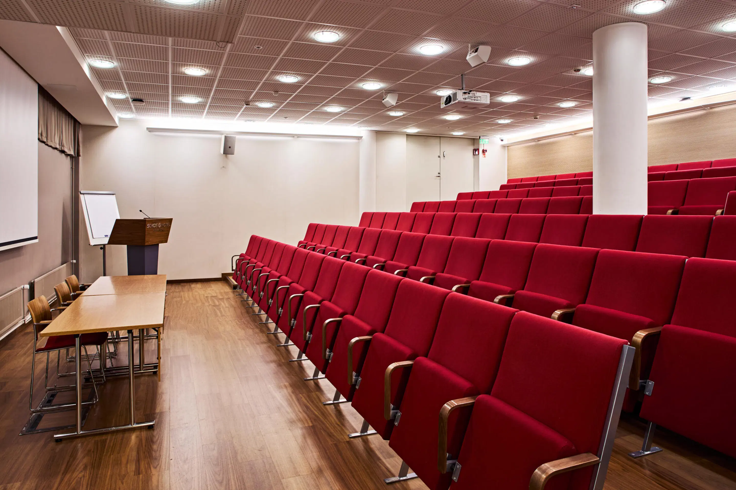 Auditorium with red seat rows, wooden speaker's table and podium at the front. Wooden flooring and white ceiling lighting.