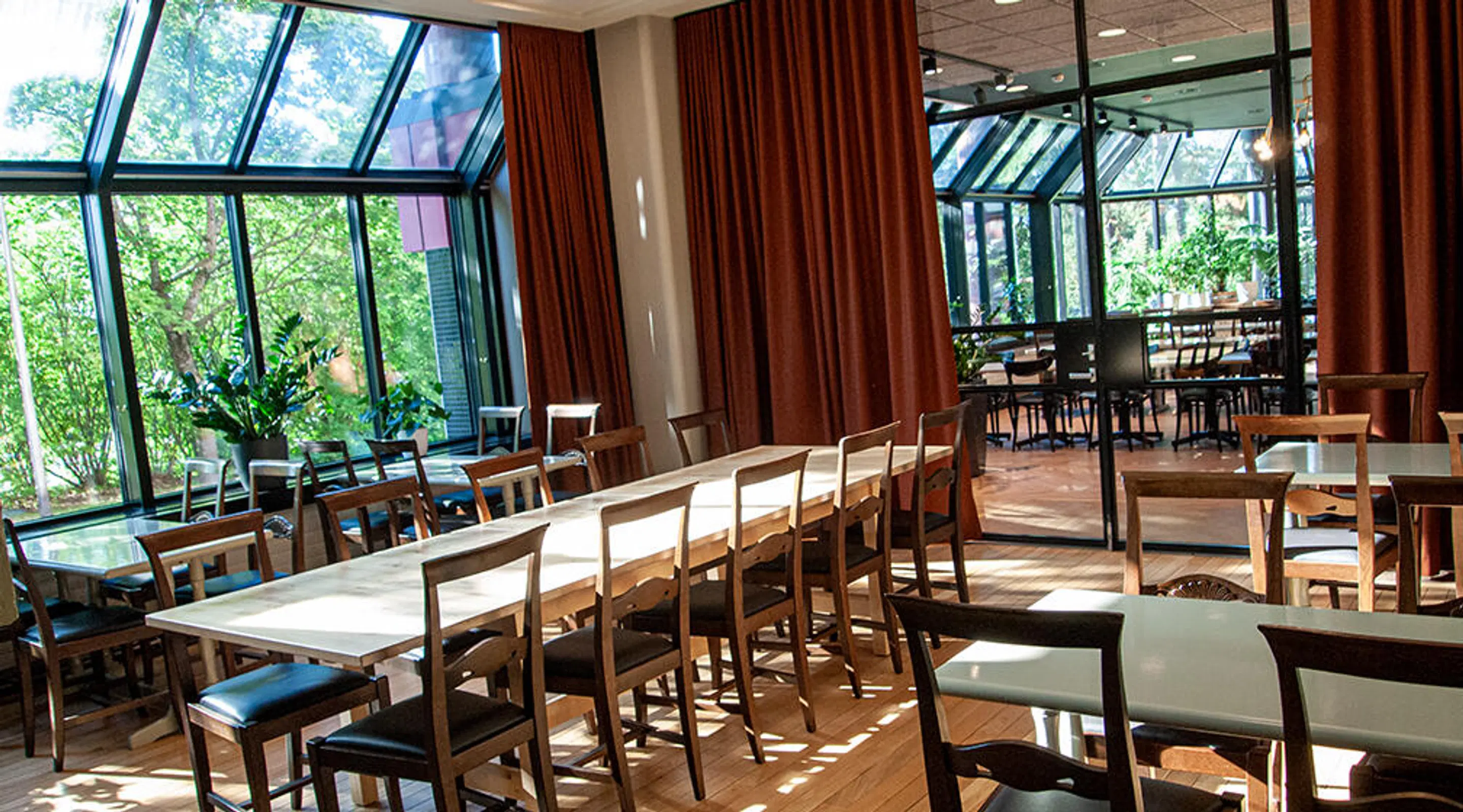 Valoisa ravintola, jossa pitkä puinen pöytä tuoleineen, lasiseiniä puutarhanäkymällä ja punaruskeilla verhoilla. Puulattia ja lisätila näkyy lasiovien takana.