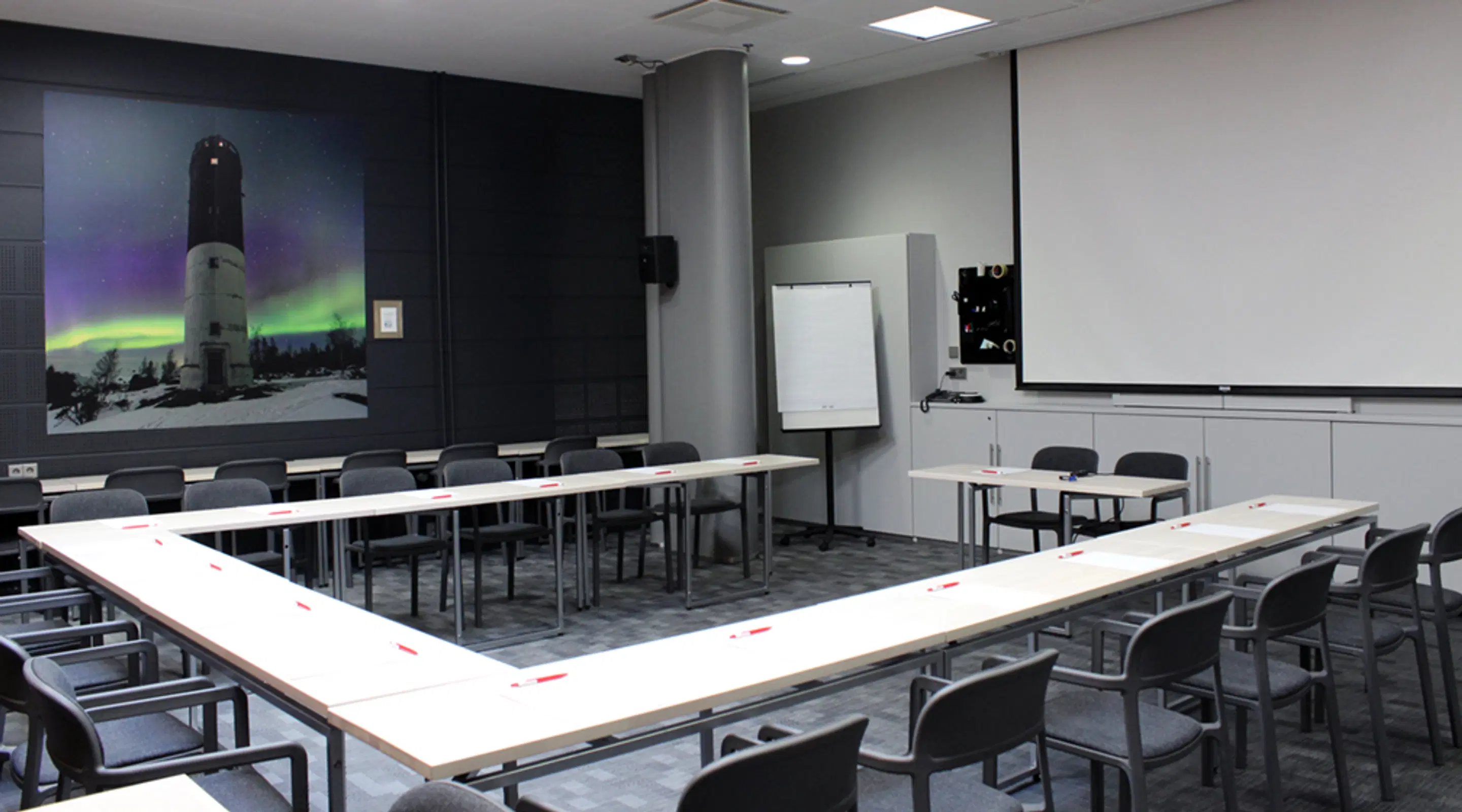 Meeting room with U-shaped table arrangement, wall featuring a lighthouse under northern lights, projector screen and whiteboard visible.