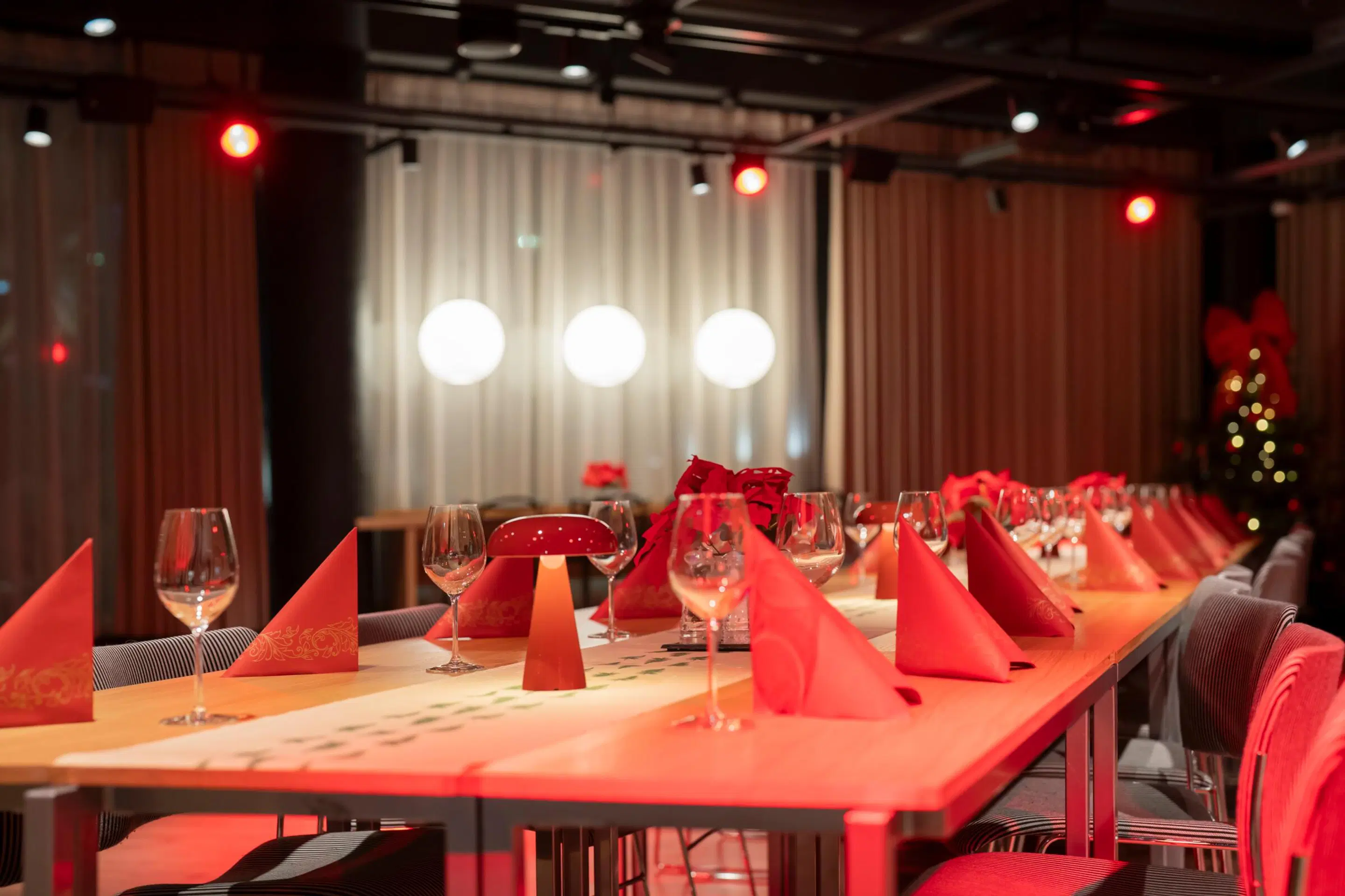 A festively set long table with red napkins and wine glasses, backdrop featuring curtains and three white lights.