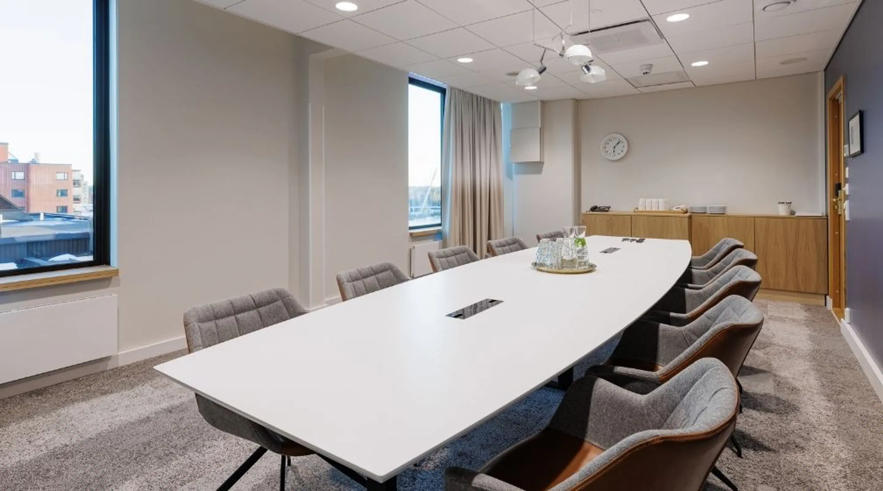 Modern meeting room with a white table, grey chairs, large windows, and wooden cabinets. The room features grey wall-to-wall carpeting and good lighting.