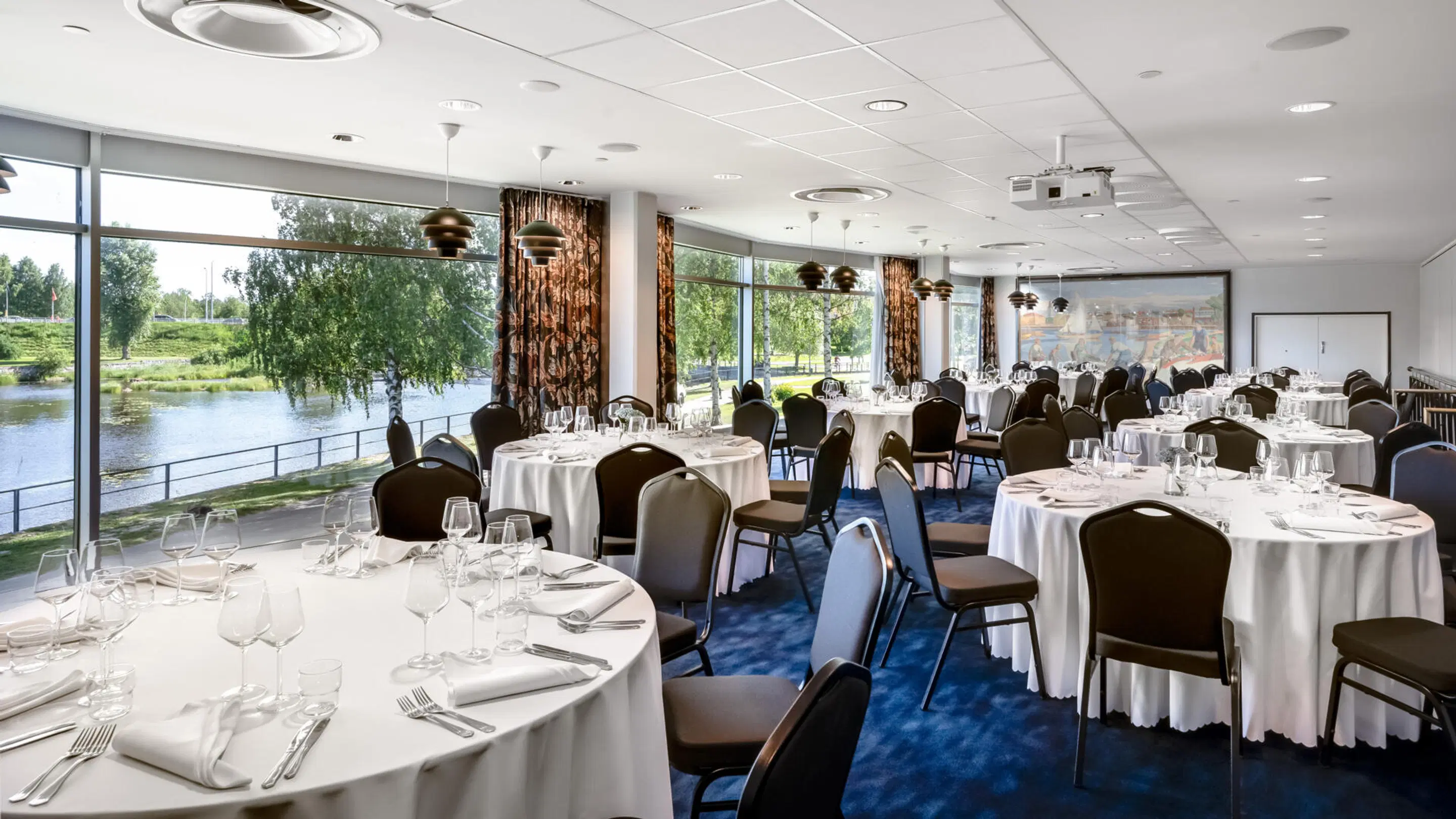 Event space with round tables covered in white tablecloths. Blue carpeted floor, large windows overlooking a lake and trees. Level floor, good lighting.