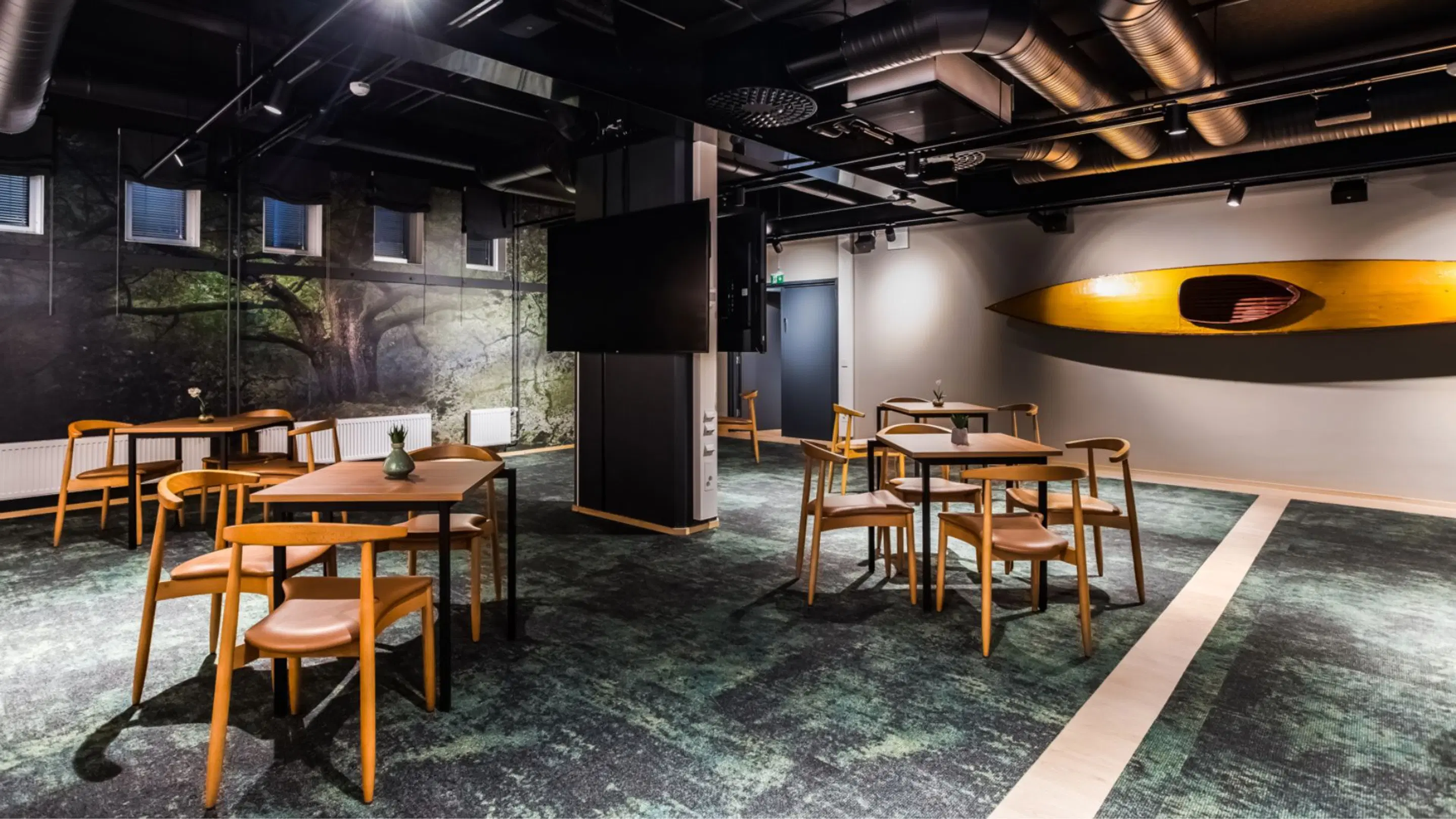 Room with small tables and wooden chairs. Green carpet on the floor, a forest mural on the wall and a yellow kayak. There is open space between the tables.