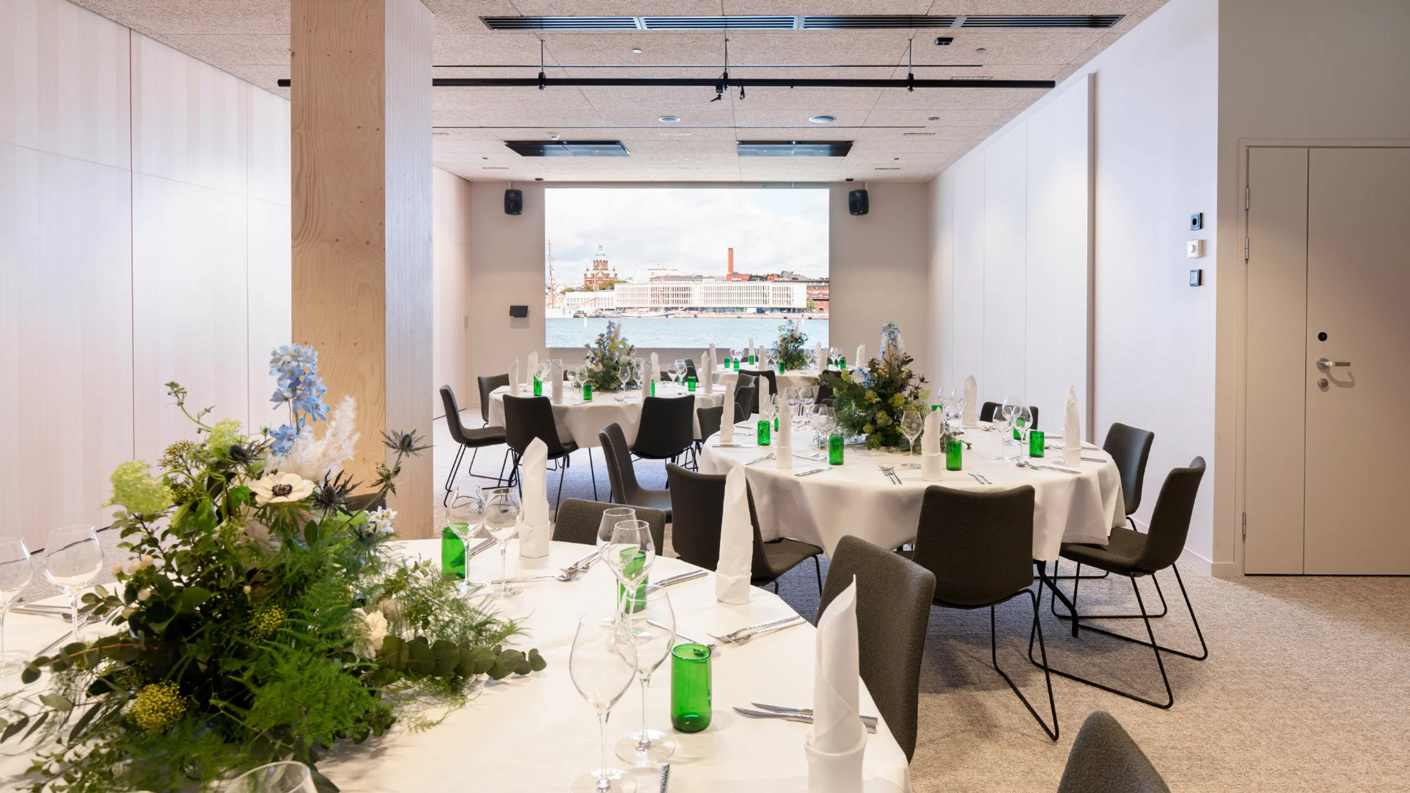 Modern meeting space with round tables, floral decorations and green glass bottles. Large screen in background reveal city view across water.