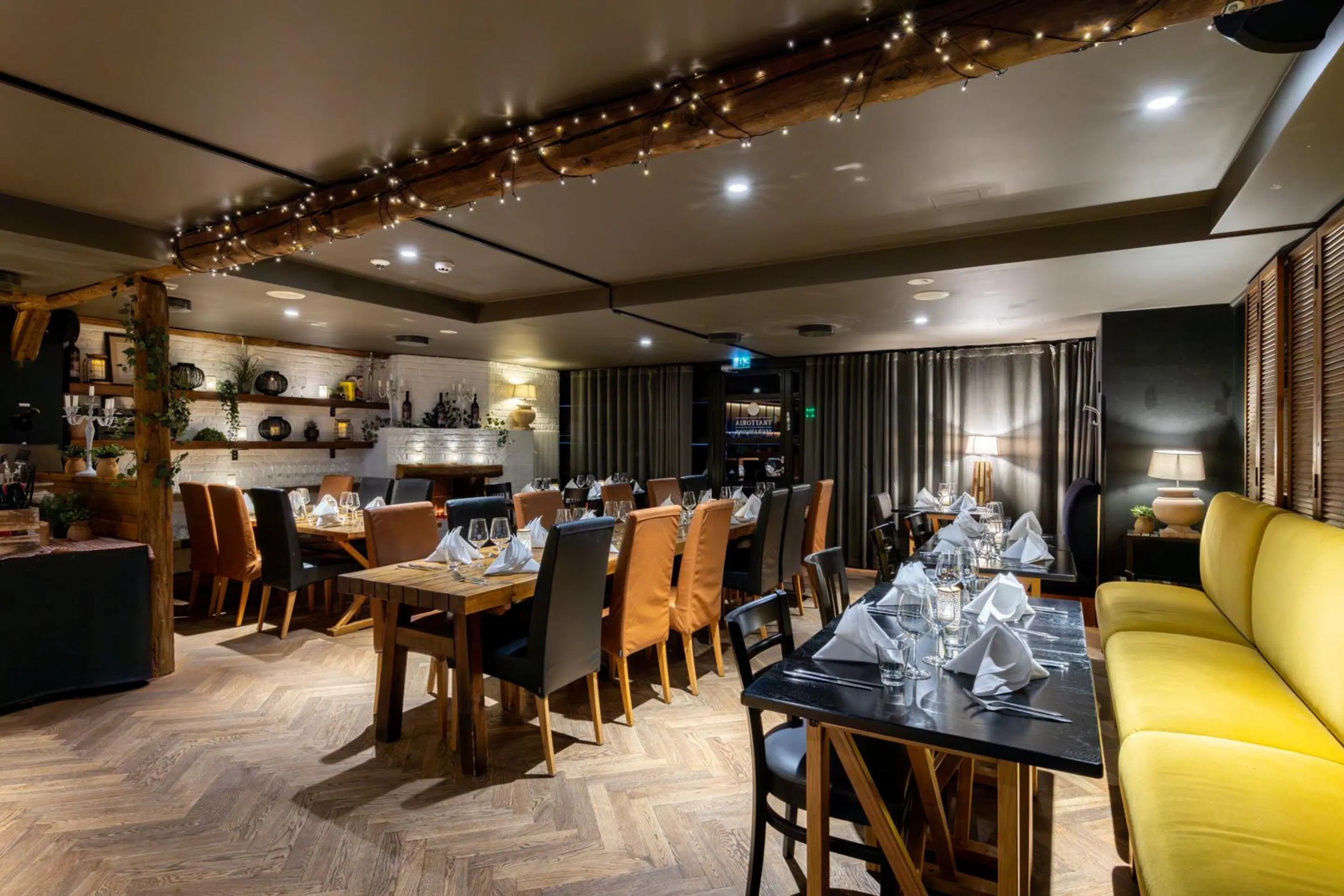 Stämningsfull restaurang med trägolv, dukade bord, träbjälkar, dekorationsbelysning och en gul soffa.