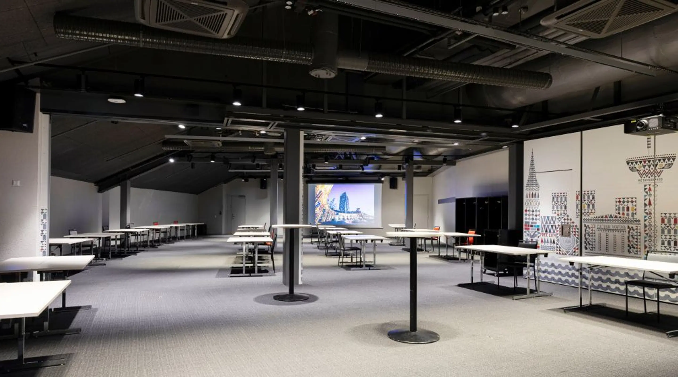Avara konferenssi- tai koulutustila, jossa pöytiä ja tuoleja, projektorinäyttö takana, näkyvissä ilmanvaihtoputkisto katossa.