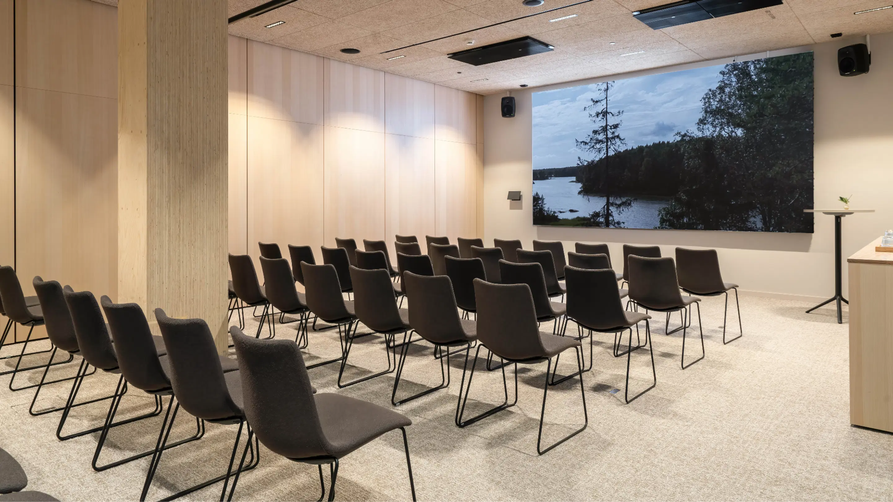 Modern auditorium with chairs arranged in rows. Large nature image of a lake landscape on the wall. Light flooring and wooden panel walls.