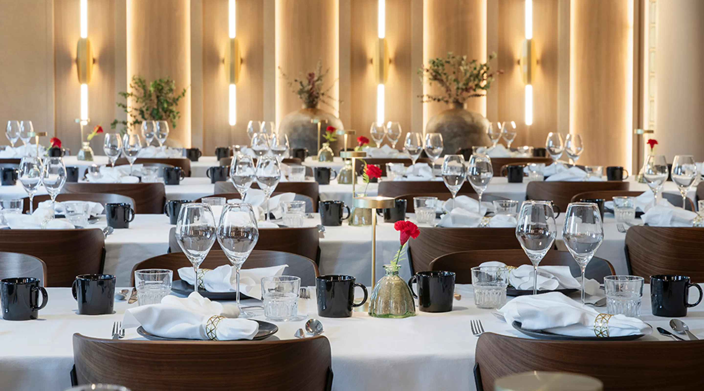 Elegant restaurangsal med bord täckta med vita dukar, röda blommor och varma väggbelysningar som skapar stämning.