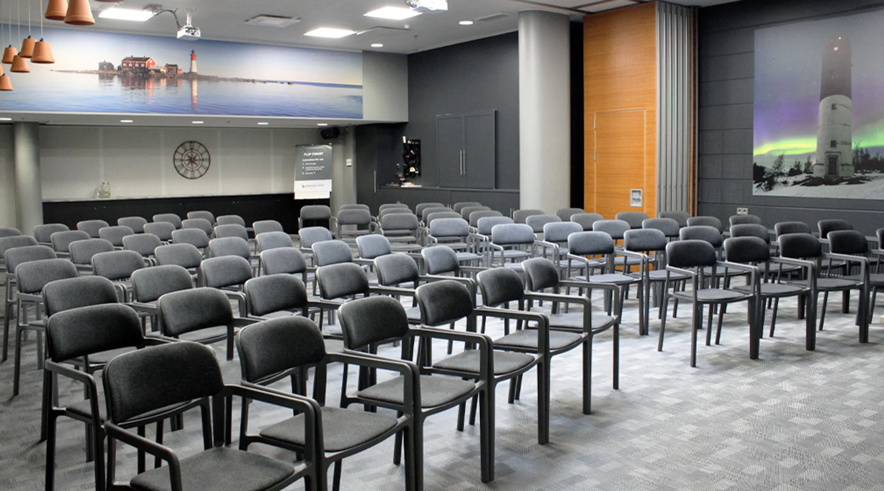 Empty auditorium with rows of grey armrest chairs. Walls feature landscape images of a lighthouse and northern lights. Level floor surface with wide aisles.