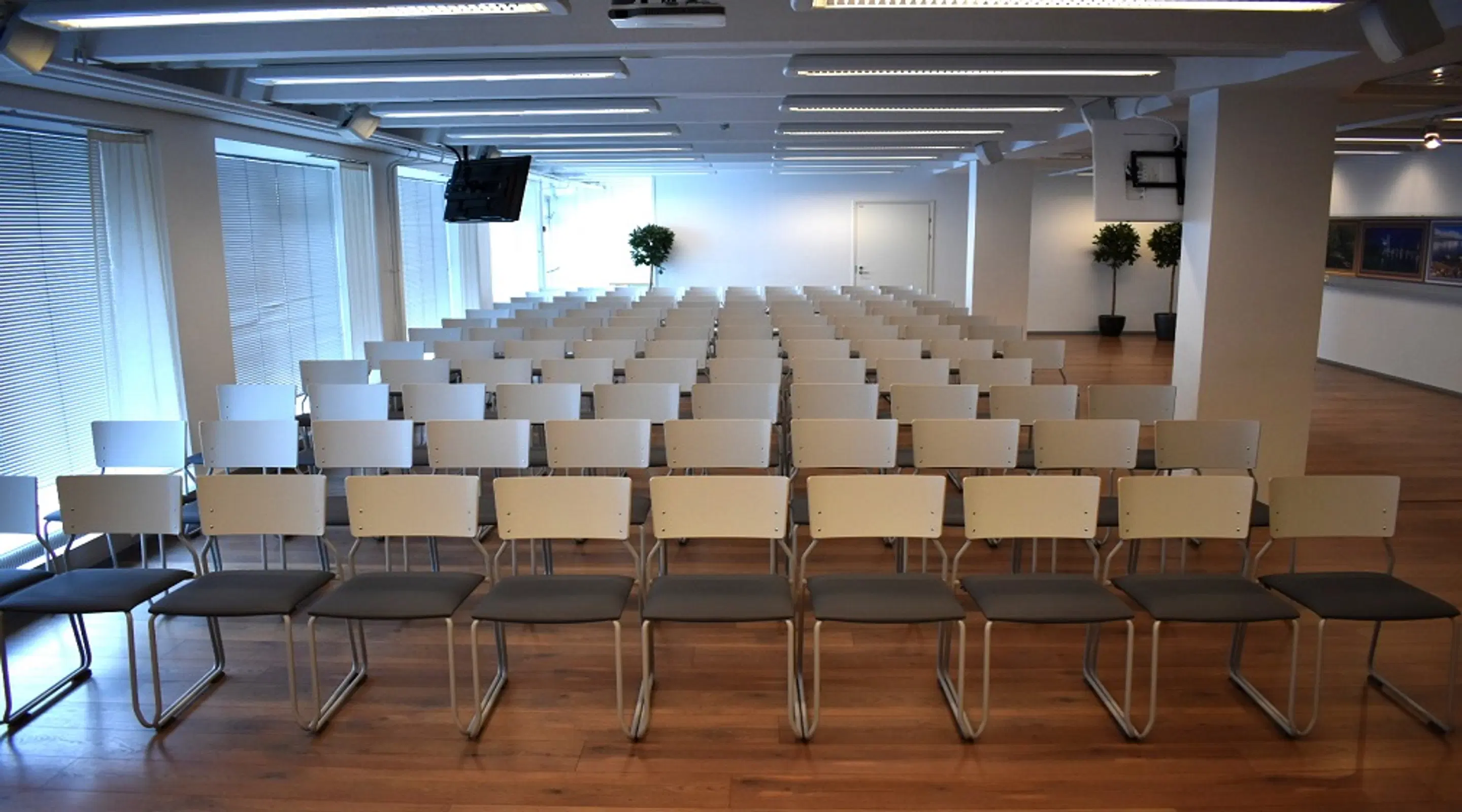 Auditorium arranged with rows of white chairs on wooden flooring. Room features large windows, a few potted plants, and a TV screen mounted on the wall.