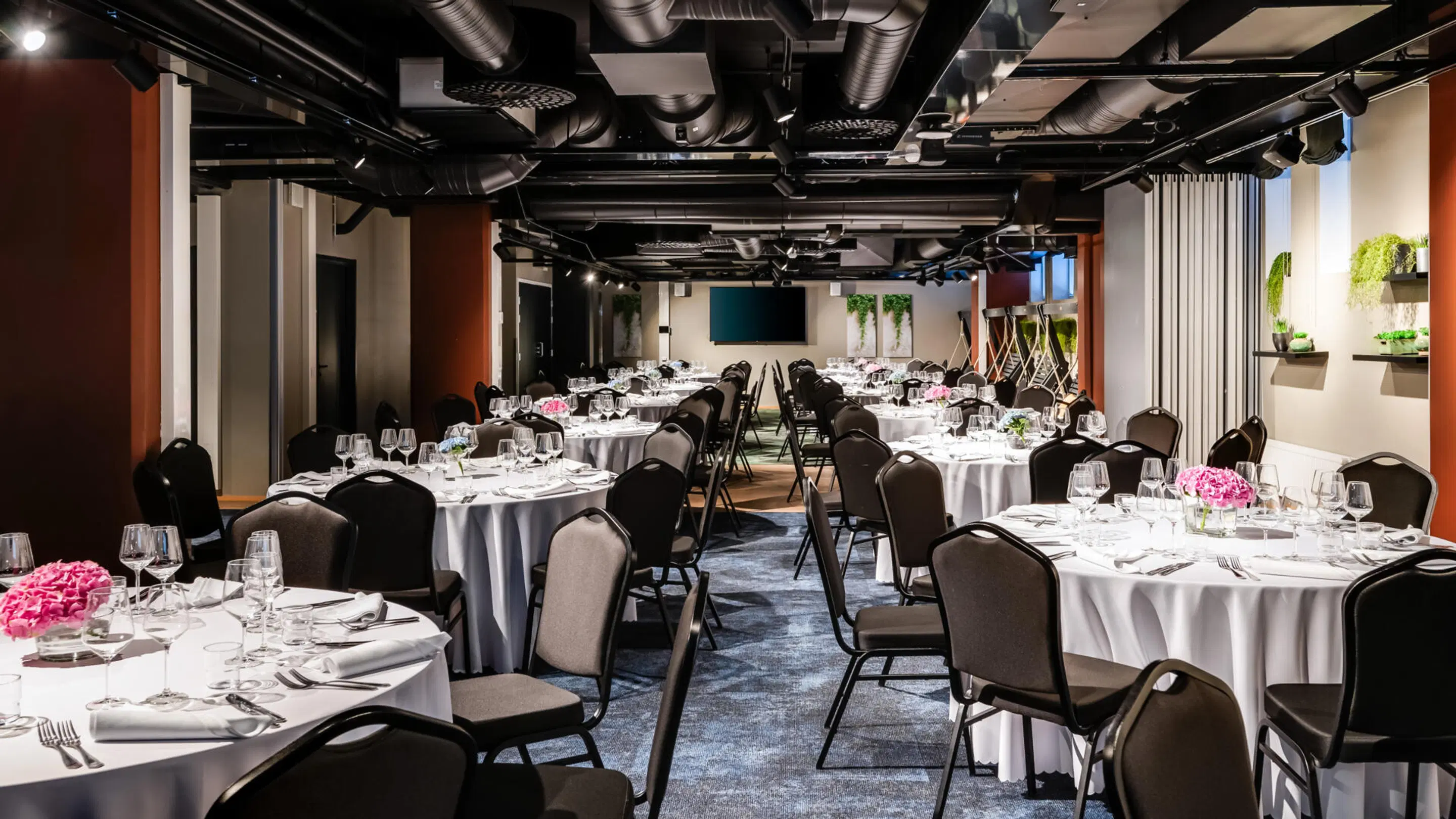 Banquet room with round tables featuring white tablecloths, wine glasses and pink flowers. Space has hard flooring and level access.