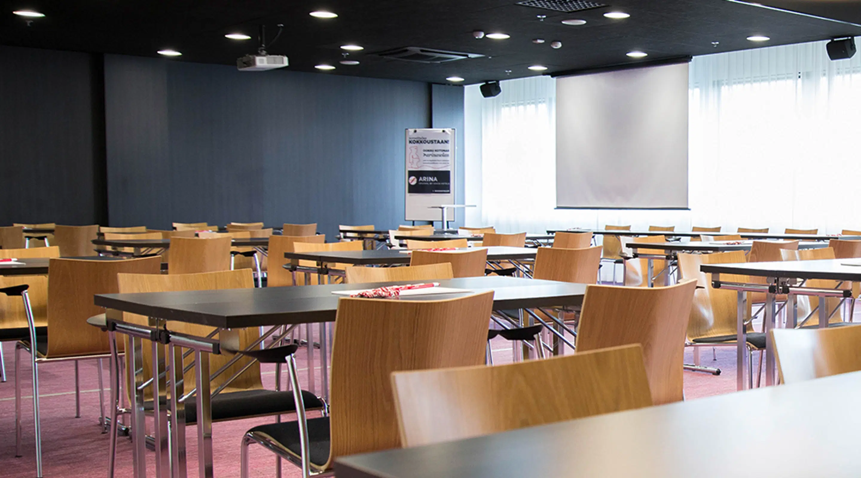 Modern meeting room with wooden chairs and work tables. Dark walls, large windows with light curtains, and ceiling-mounted projector.