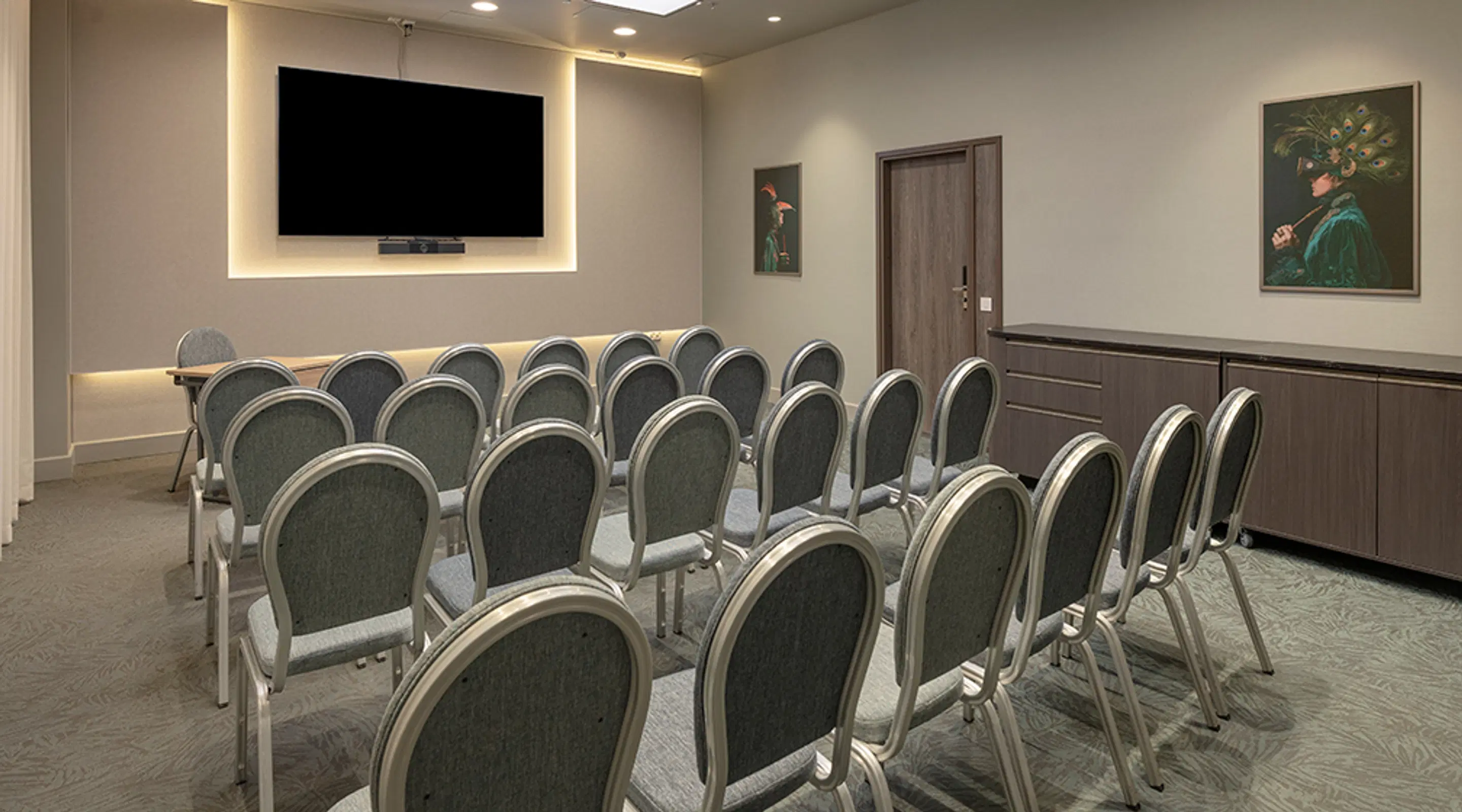 Small meeting room with rows of grey upholstered chairs and a large wall-mounted screen. The floor has patterned carpeting and the lighting is soft.