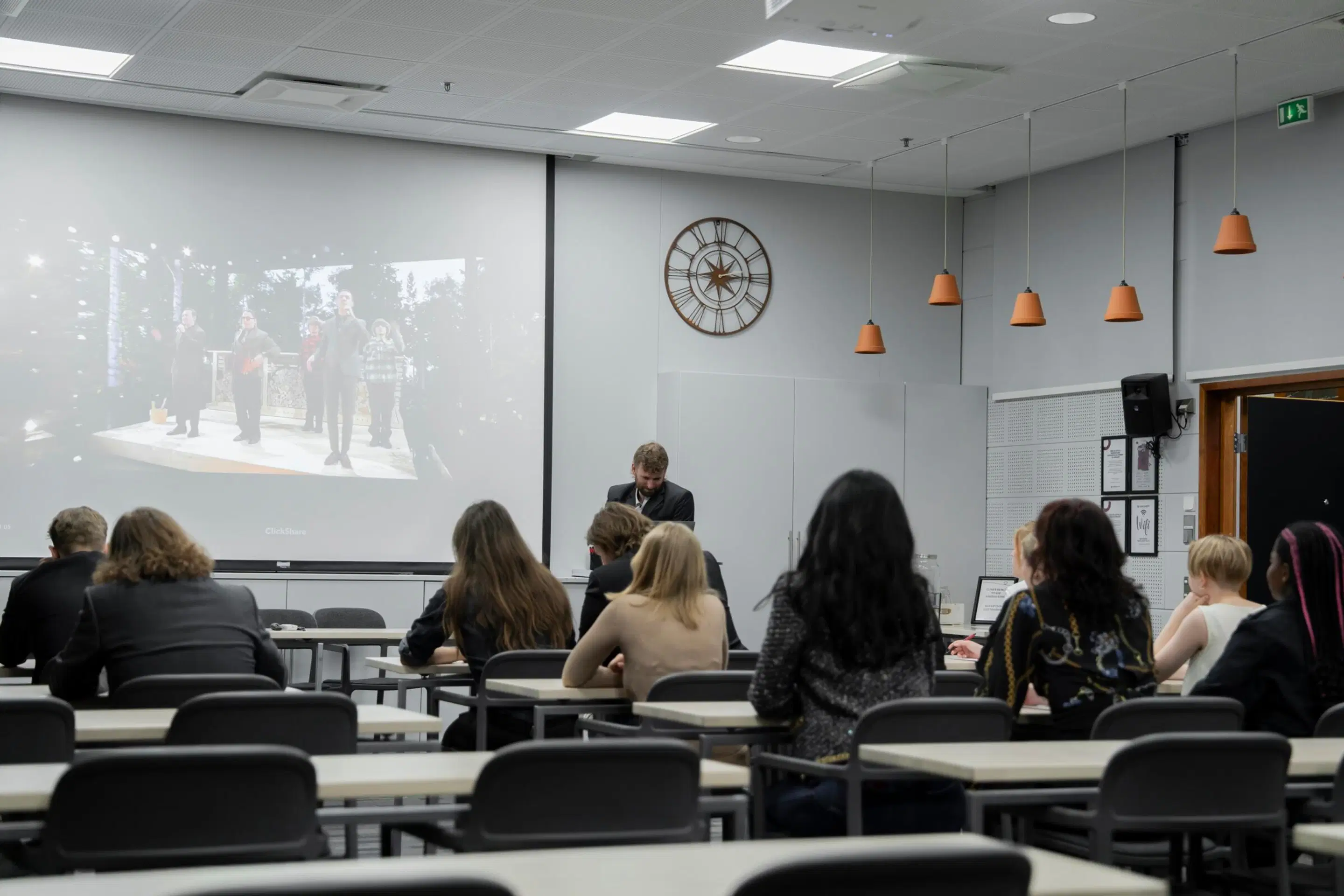 Attendees in a modern classroom watching a presentation. The white room features a projector screen, wall clock, and orange pendant lights.