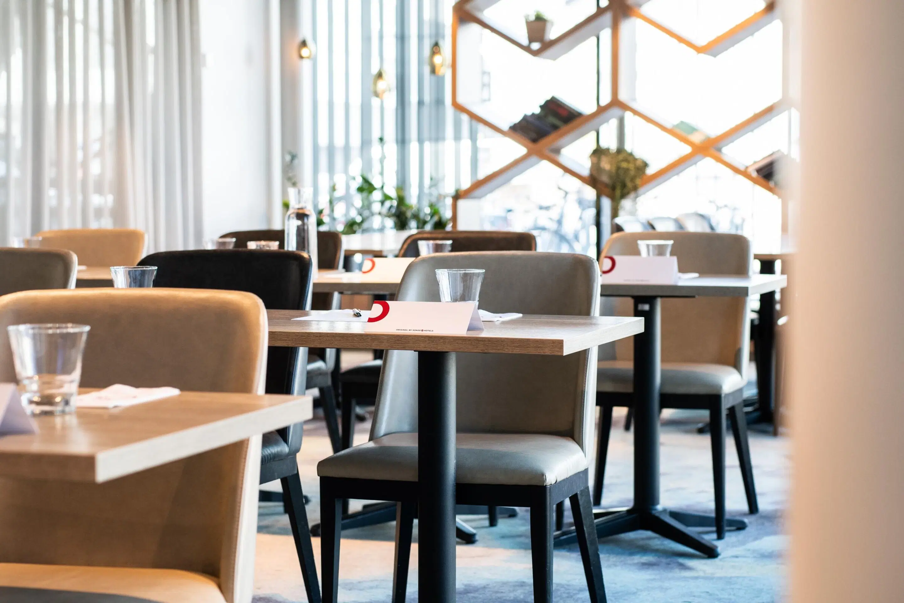 Restoran laudade ja toolidega pehmel vaibakattega. Avar käigutee laudade vahel. Suur geomeetriline riiul seinal.