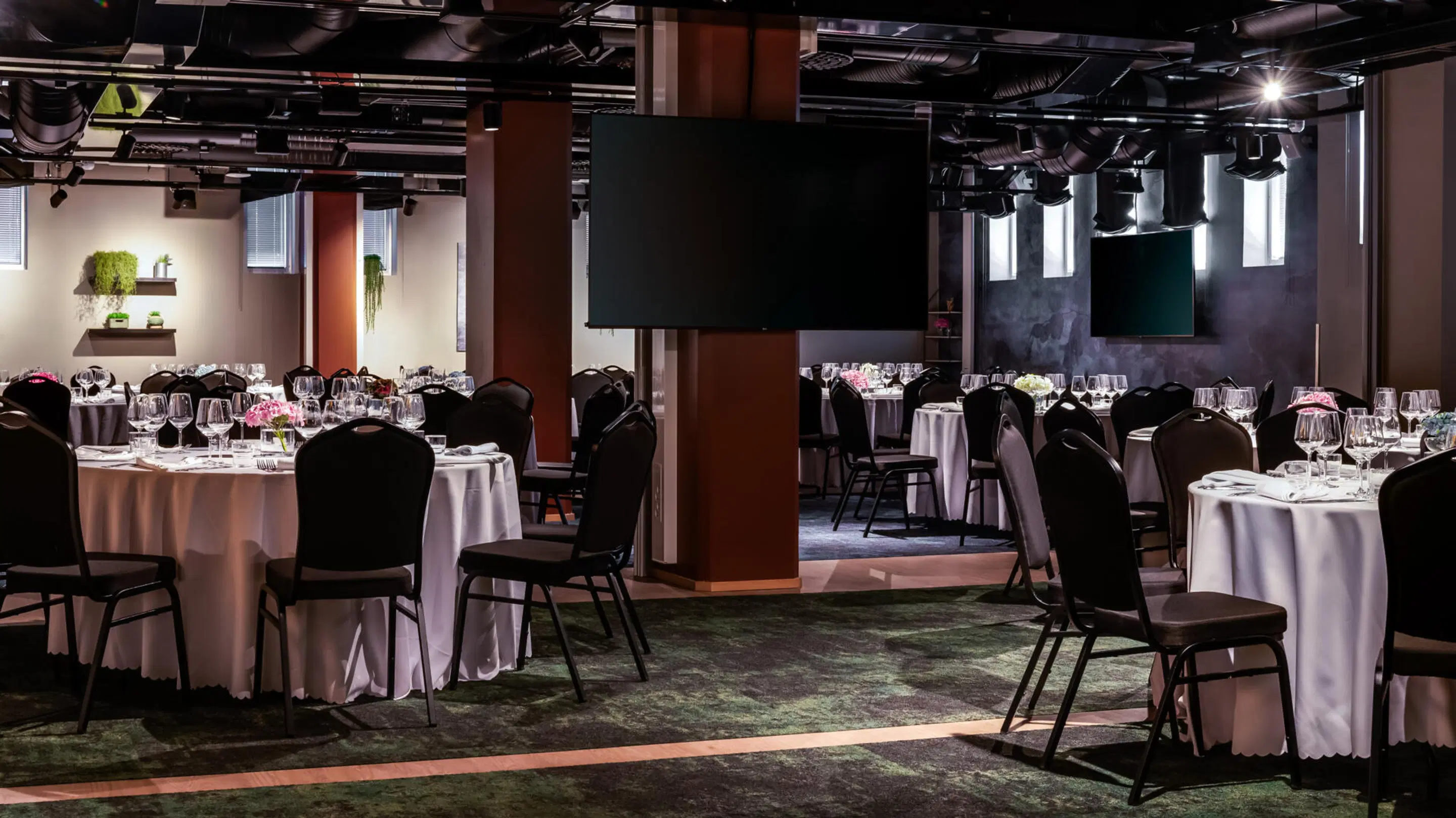 Modern banquet room with round tables and black chairs. White tablecloths, wine glasses, displays and green plants in background. Level floor and open space.