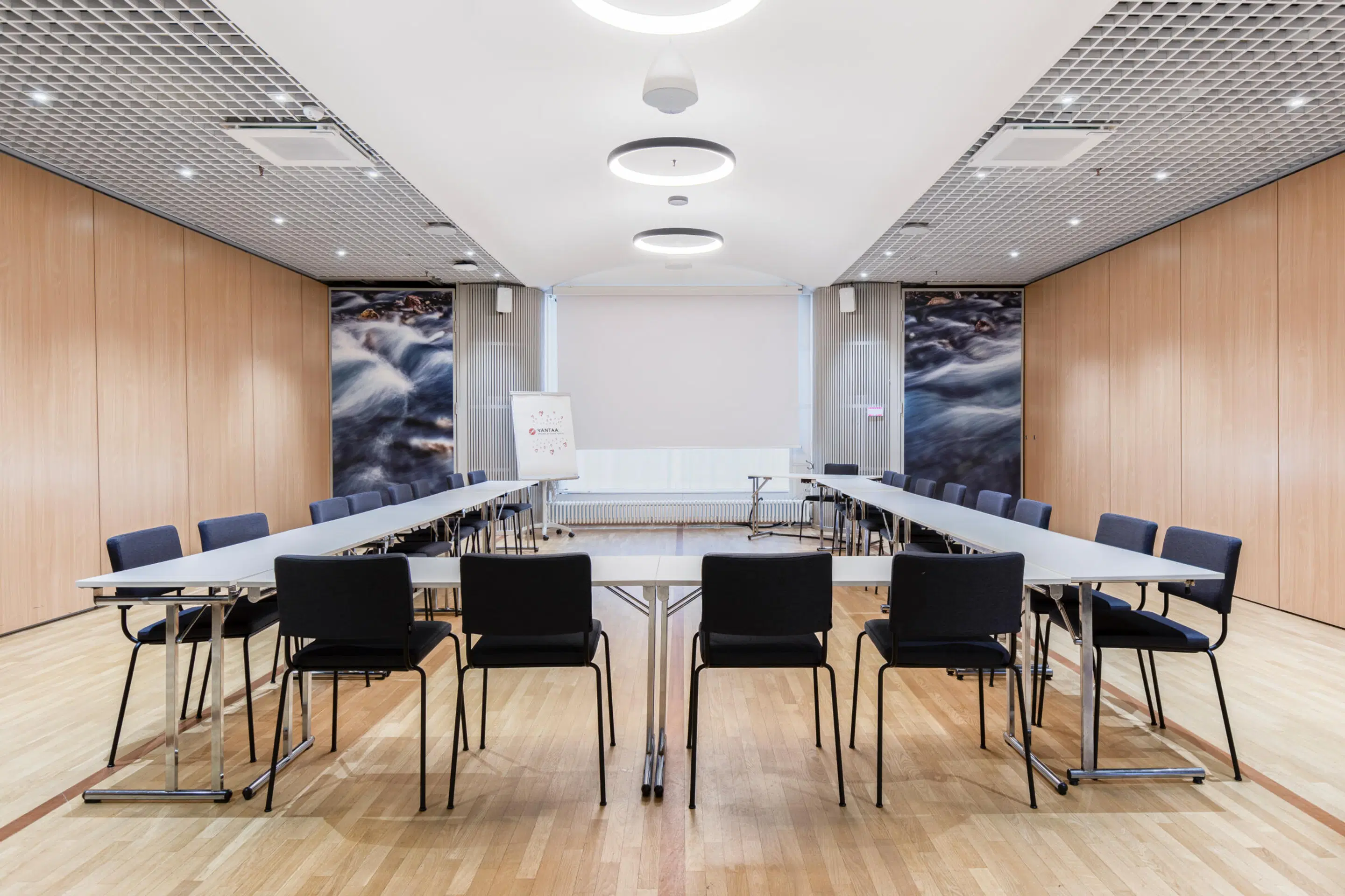 Modern meeting room with U-shaped table arrangement, wooden wall panels, water-themed artwork, light wooden flooring and stylish ceiling lights.