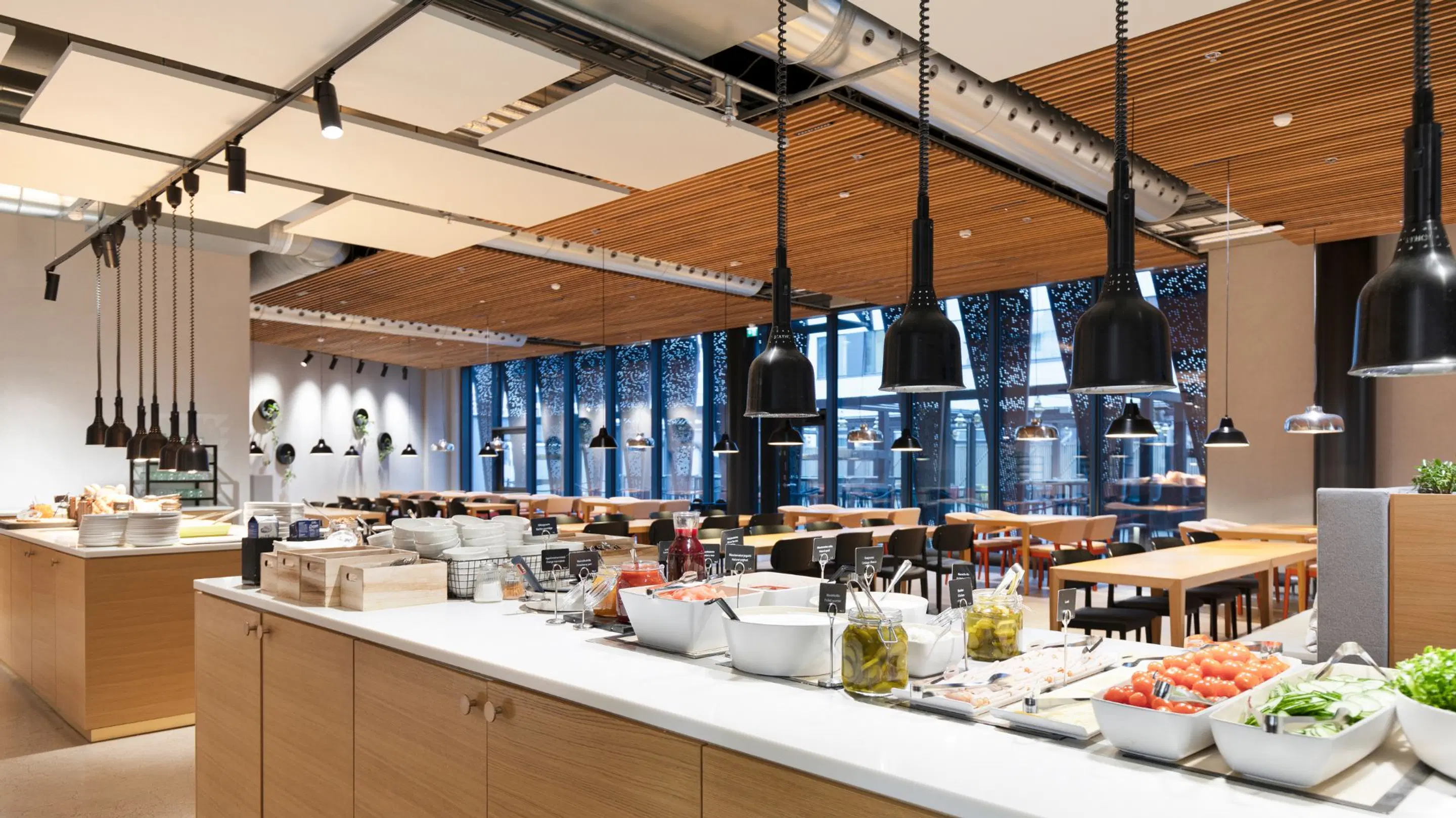 Modern restaurant with buffet counter in foreground. Spacious dining area features wooden ceiling, black pendant lights and large windows. Floor has no carpets.