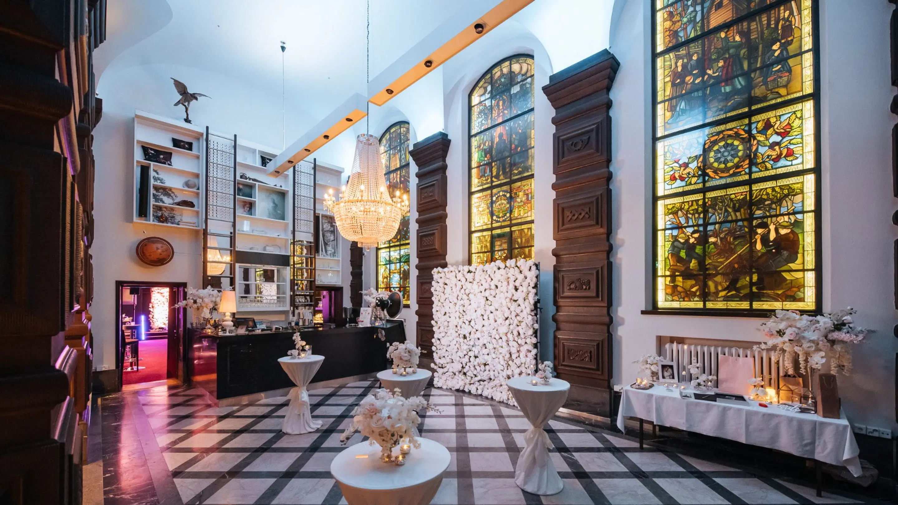 Elegant hall with tall stained glass windows, crystal chandelier and marble floor. White floral arrangements on tables and floral installation. Space has level flooring and wide pathways.
