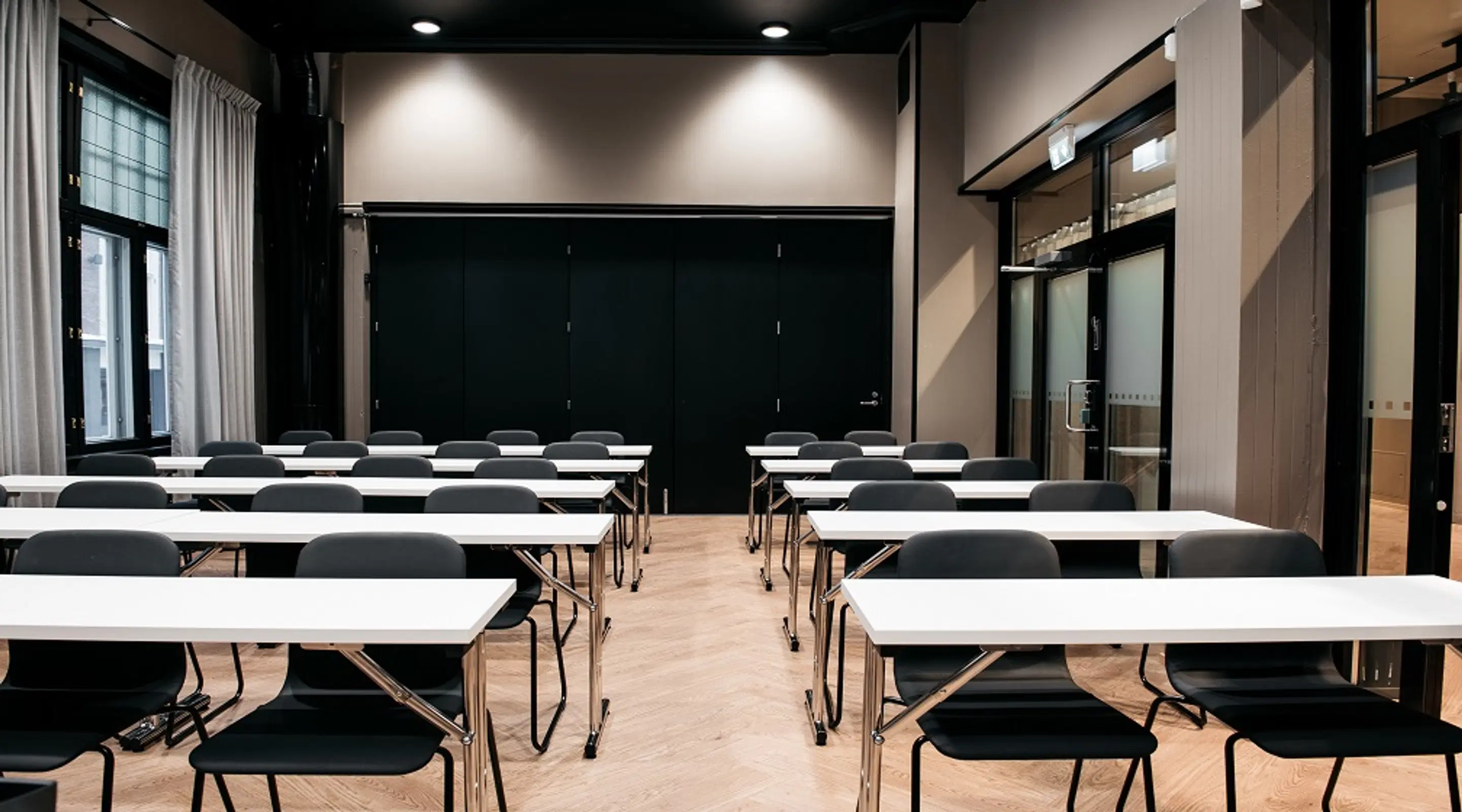 Modern meeting room with rows of white tables and black chairs, wooden flooring, dark folding doors, and windows with light curtains.