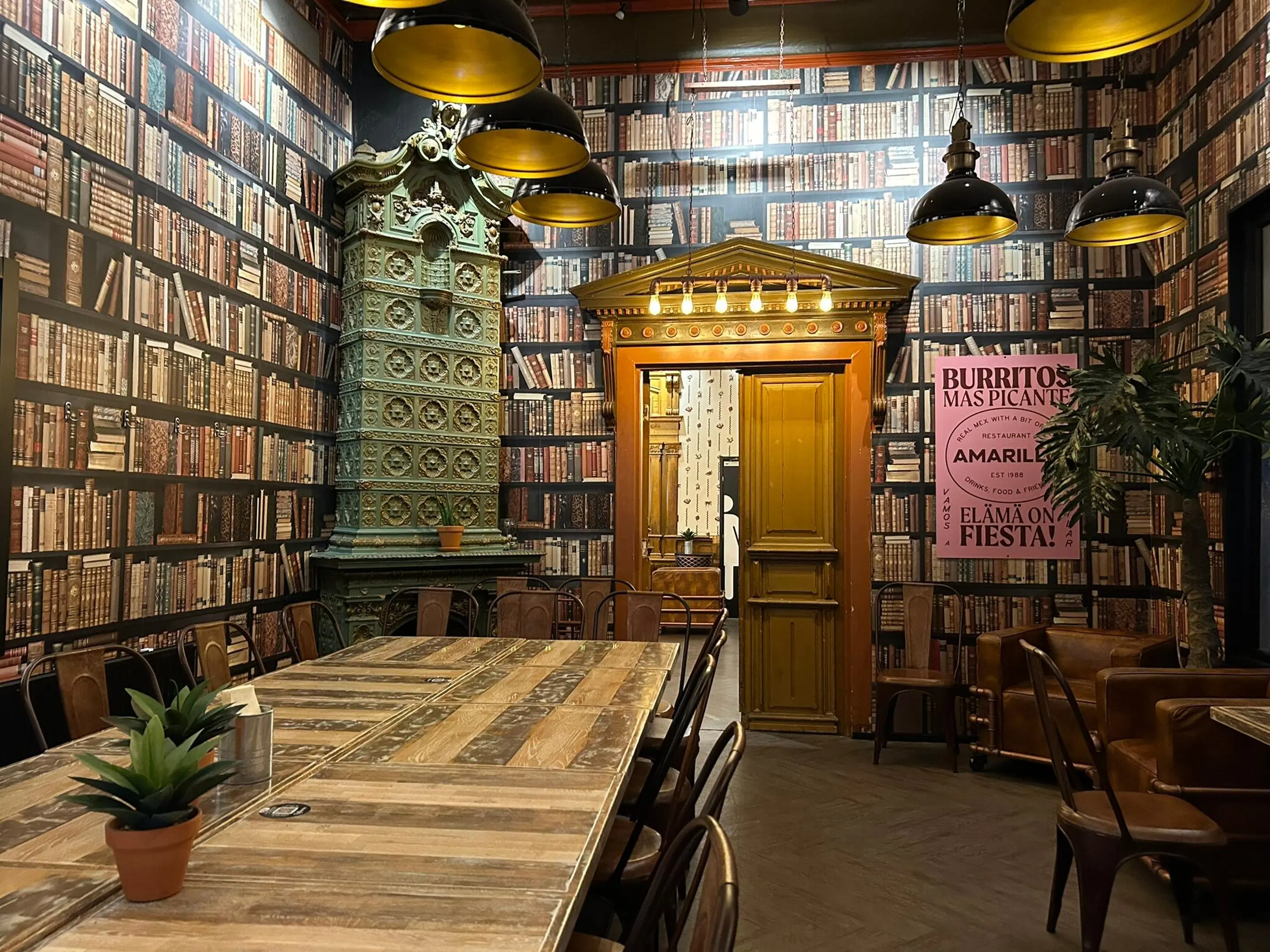 Restaurant with library wallpaper, green tile stove, wooden door and long table. Black pendant lights with gold interior hang from ceiling.