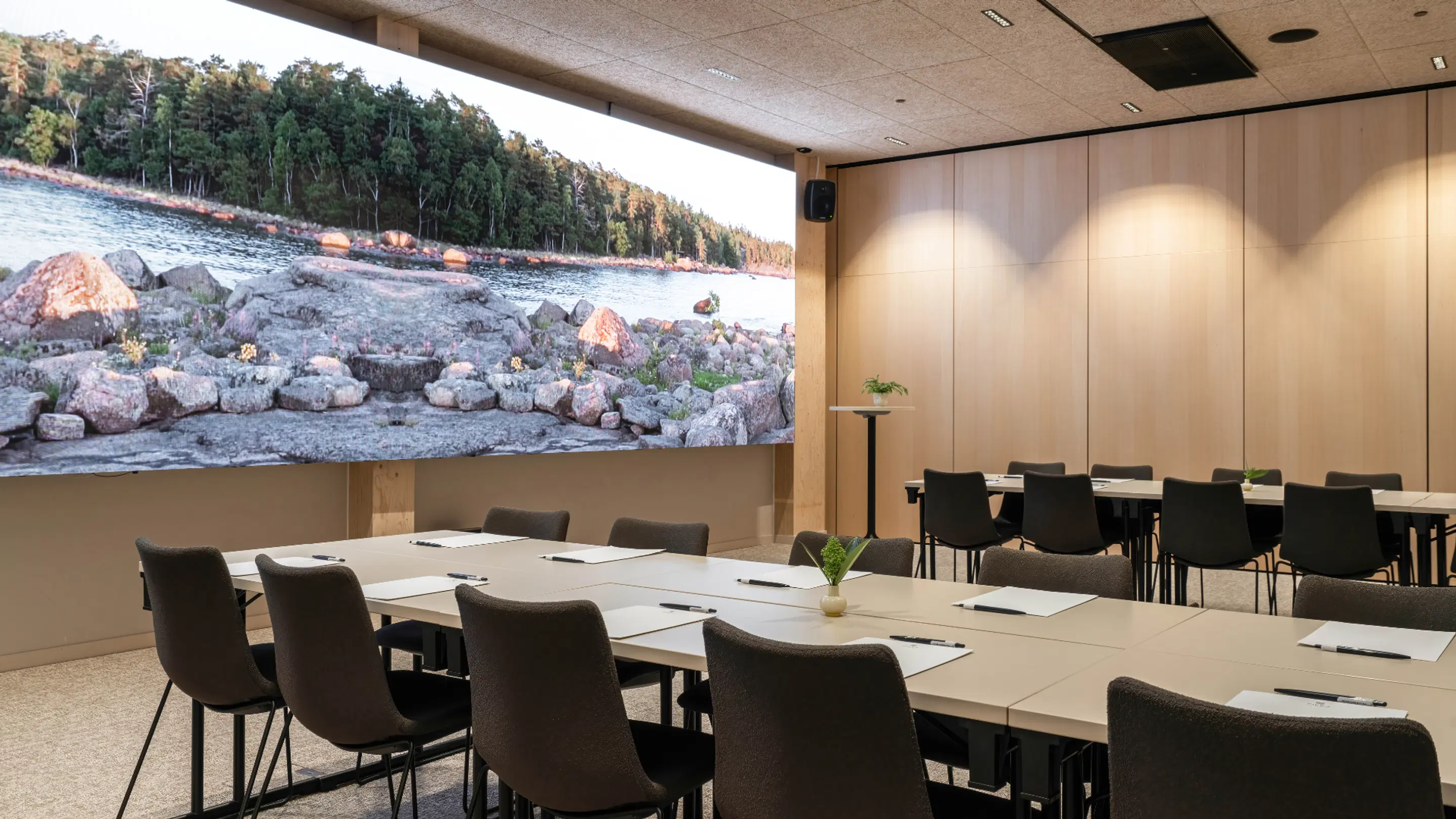 Modern conference room with light wood paneling and large wall display showing a lakeside scene with rocky shore. Tables set with notepads and pens.
