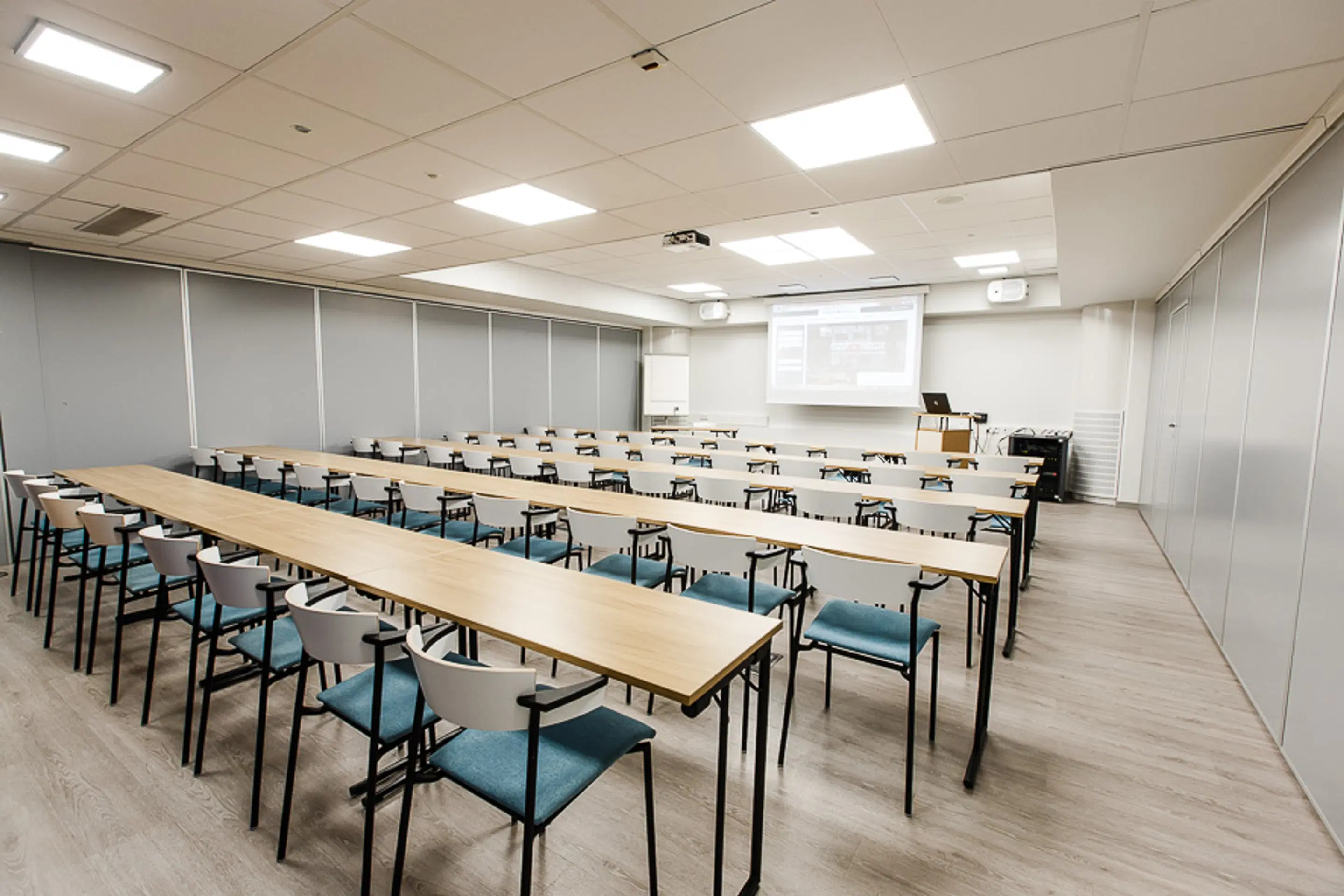 Modern classroom with long wooden tables and blue chairs. Light wooden floor and projector at the front. Spacious room with bright lighting.