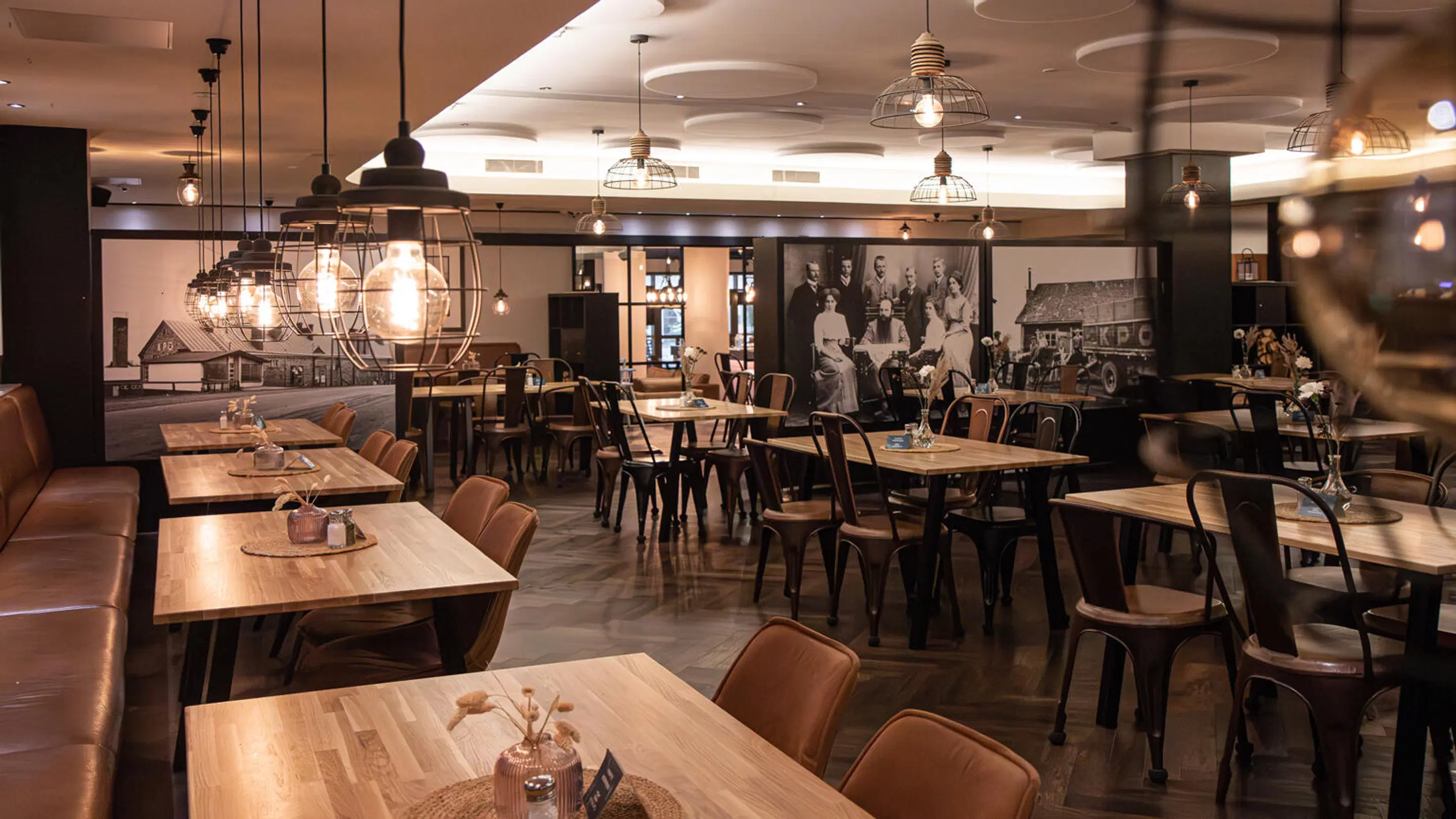 Restaurant interior with wooden tables, brown chairs and leather sofas. Vintage-style lighting fixtures and a wall with historical photographs.