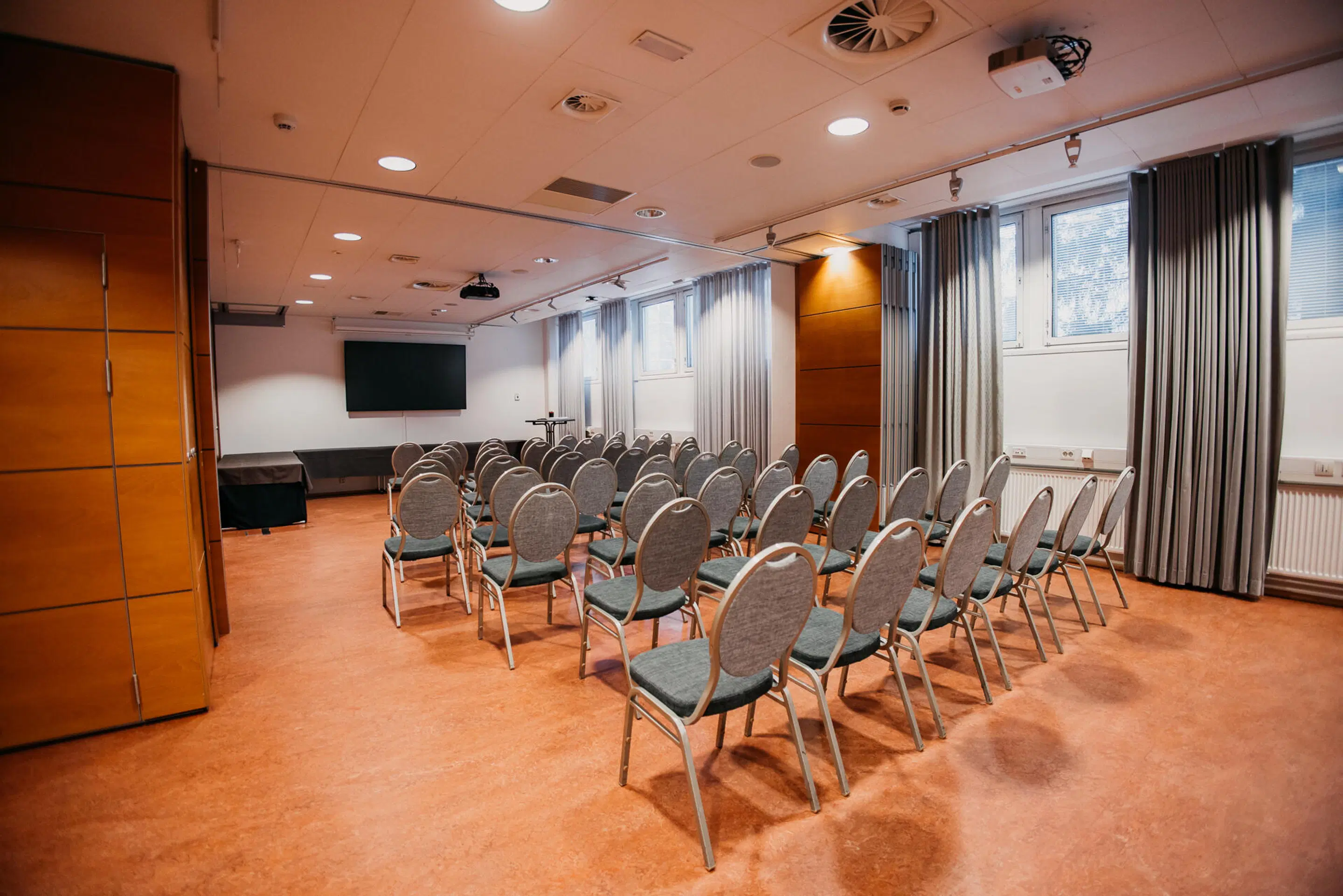 Konferensrum med grå stolar i rader, projektor, skärm och träpanelerade väggar. Plant golv och god belysning.