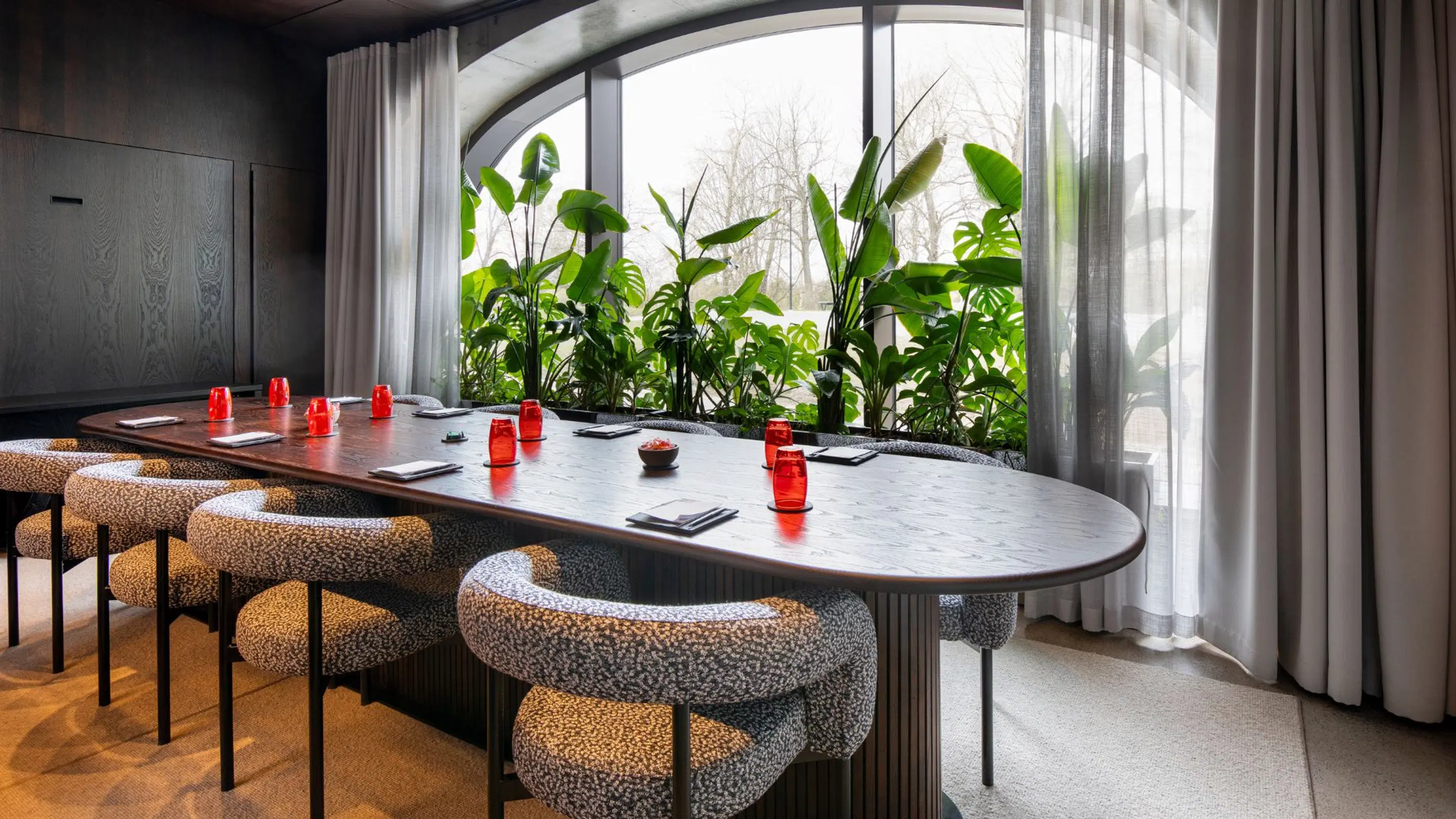 Modern meeting room with long wooden table and leopard print chairs. Red glasses on table, large windows with green plants behind.