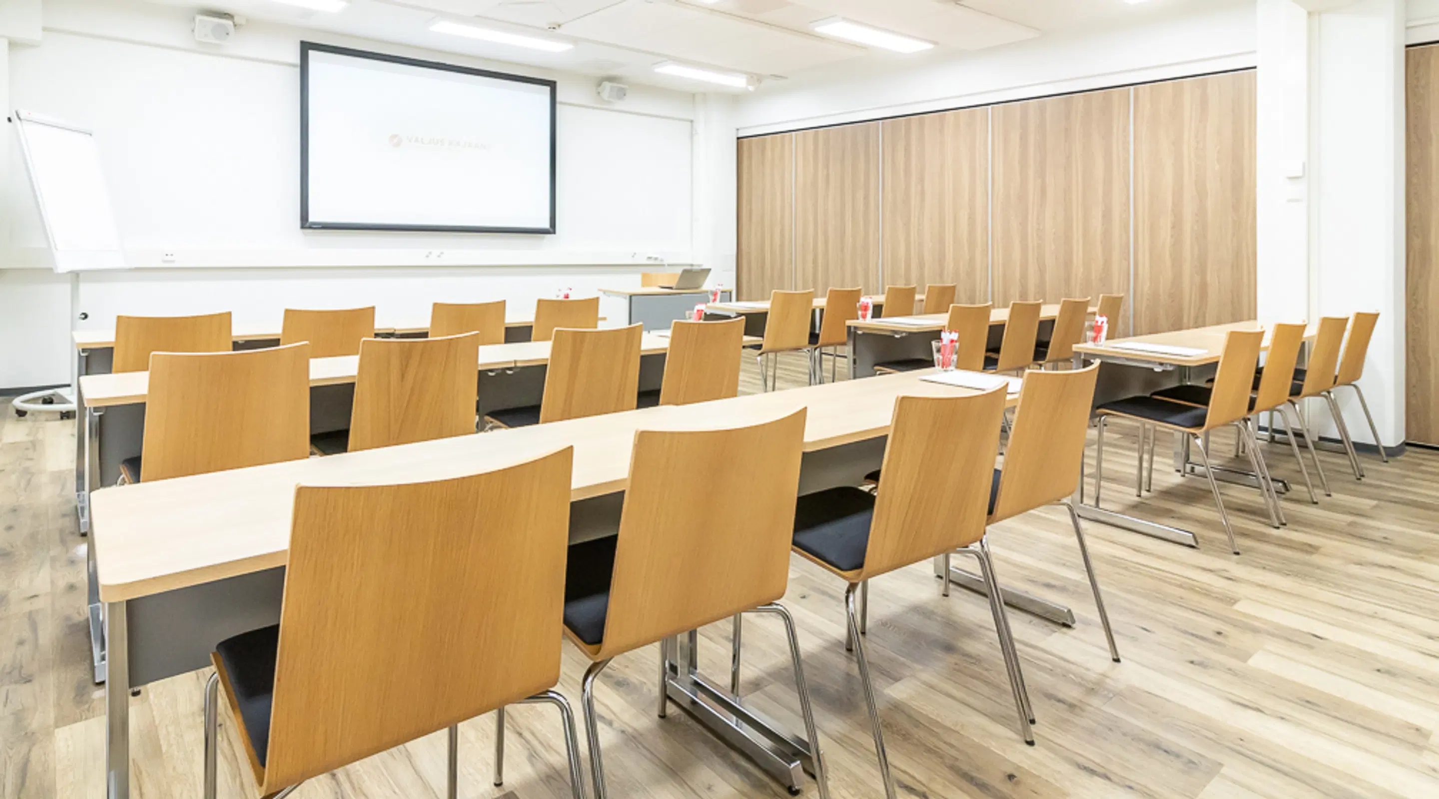 Conference room with rows of light wooden tables and chairs, a projector screen, and light wooden panels on the wall.