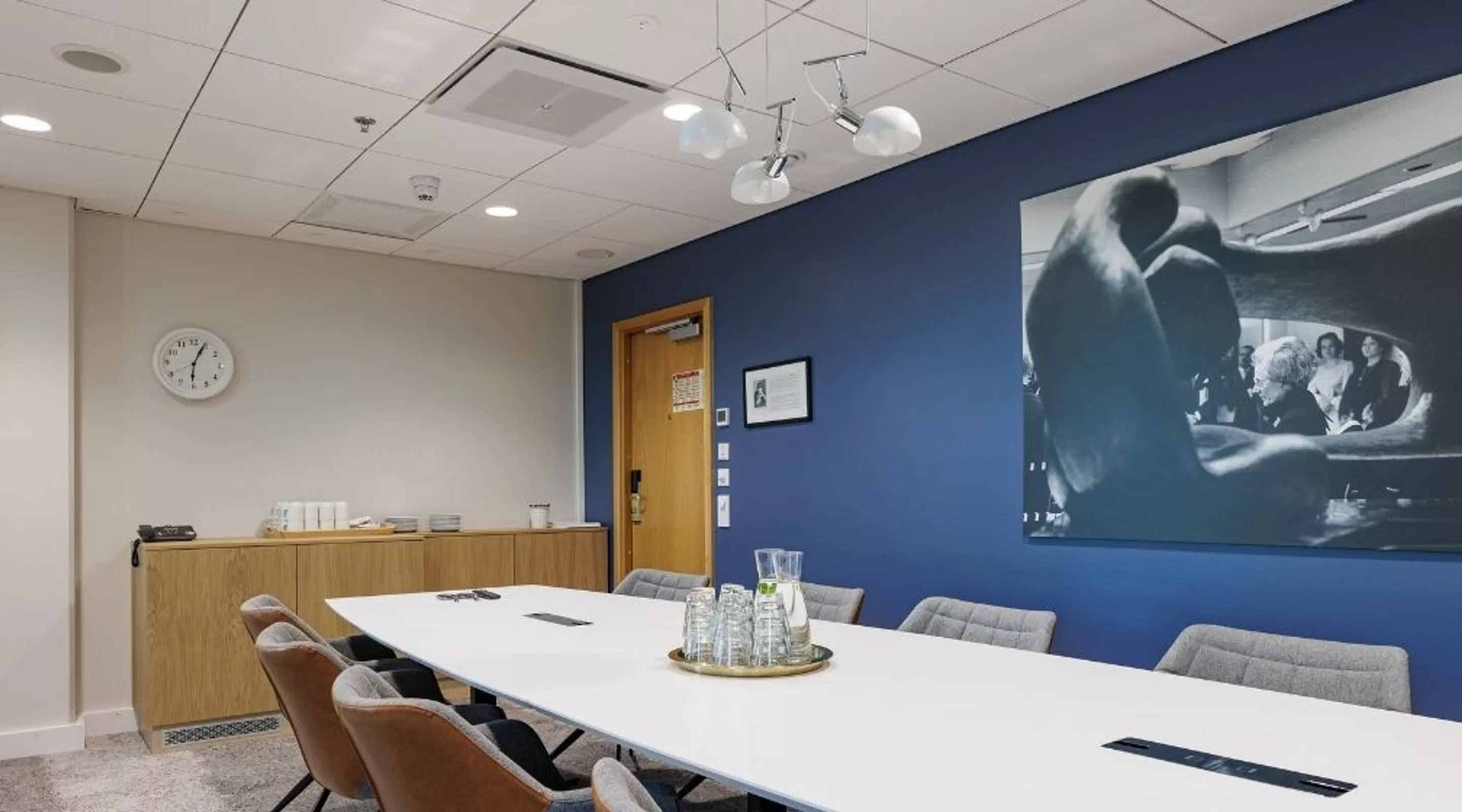 Modern meeting room with white table, gray chairs, blue accent wall and wooden door. Water carafes on tray at center of table.