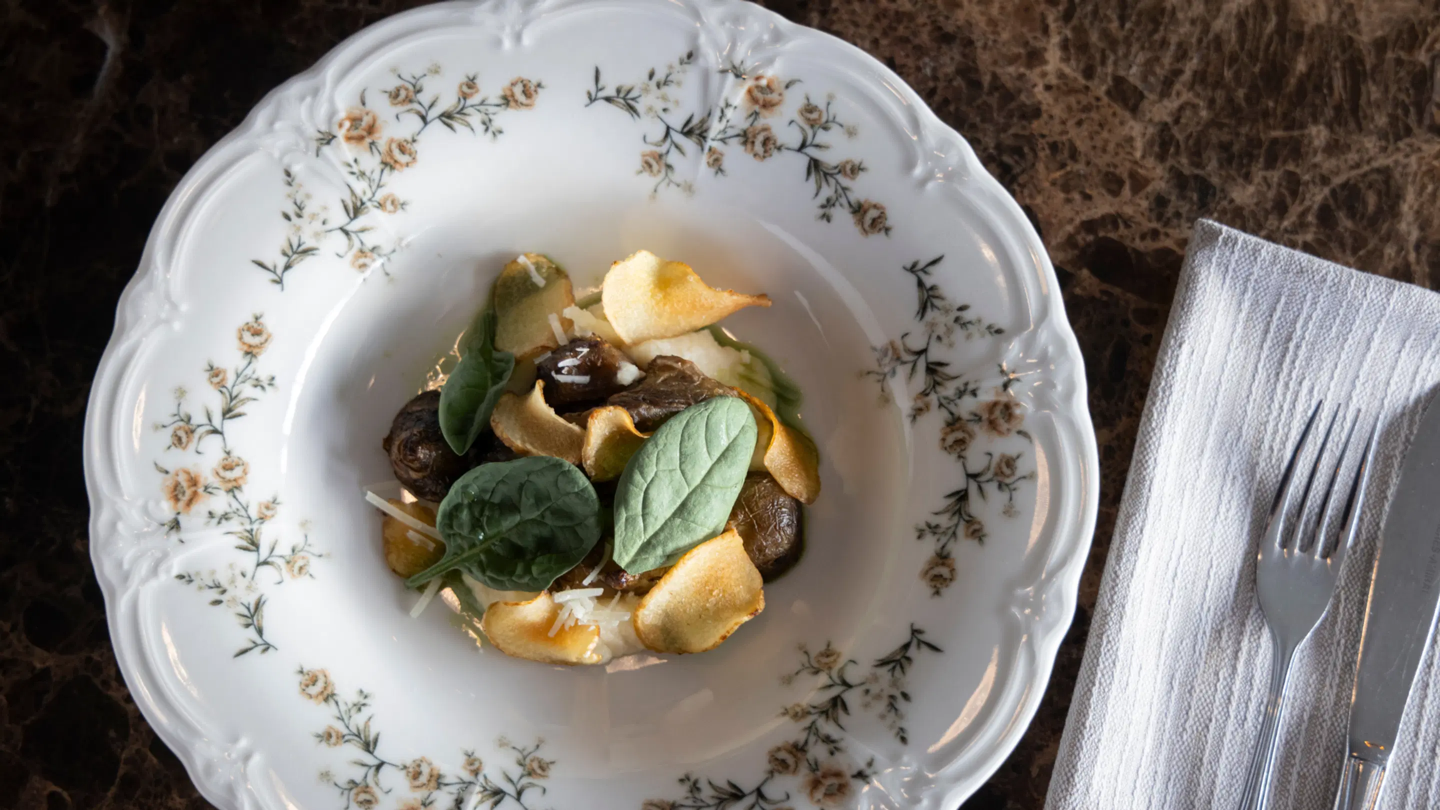 Decorative porcelain plate with a mushroom dish, spinach leaves, and potato crisps. A white napkin and fork sit alongside.