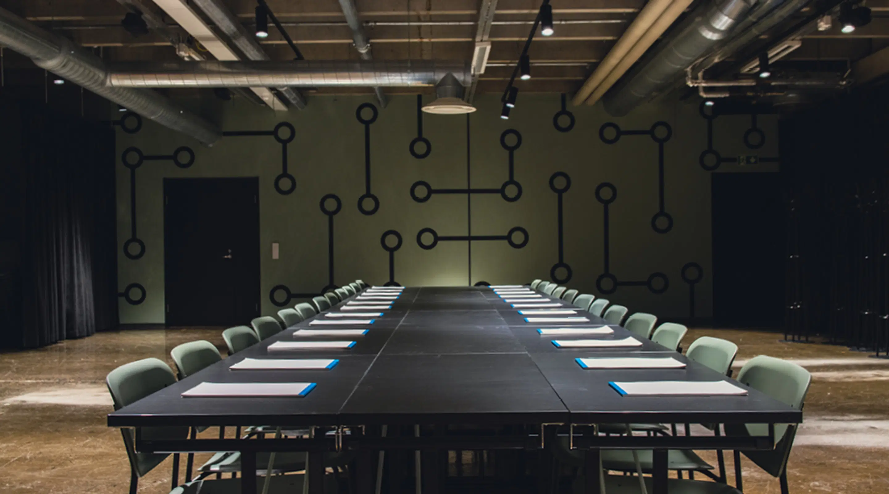 Conference room with a long dark table and light green chairs. Green wall with circuit pattern in background.