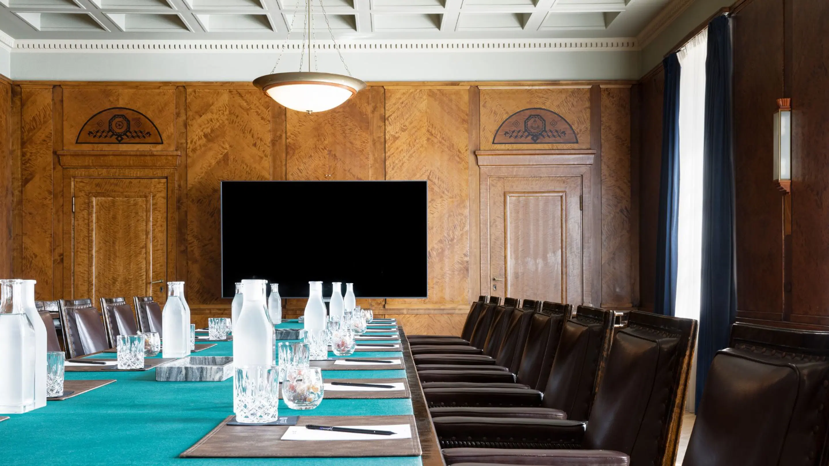 Elegant meeting room with wooden paneling, long table with turquoise runner, leather chairs and large display screen on wall.