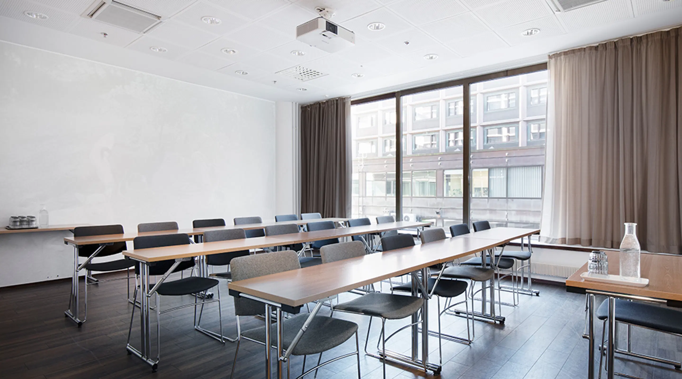 Modern meeting room with long tables and grey chairs arranged in rows. Large windows, ceiling-mounted projector and dark wooden floor.