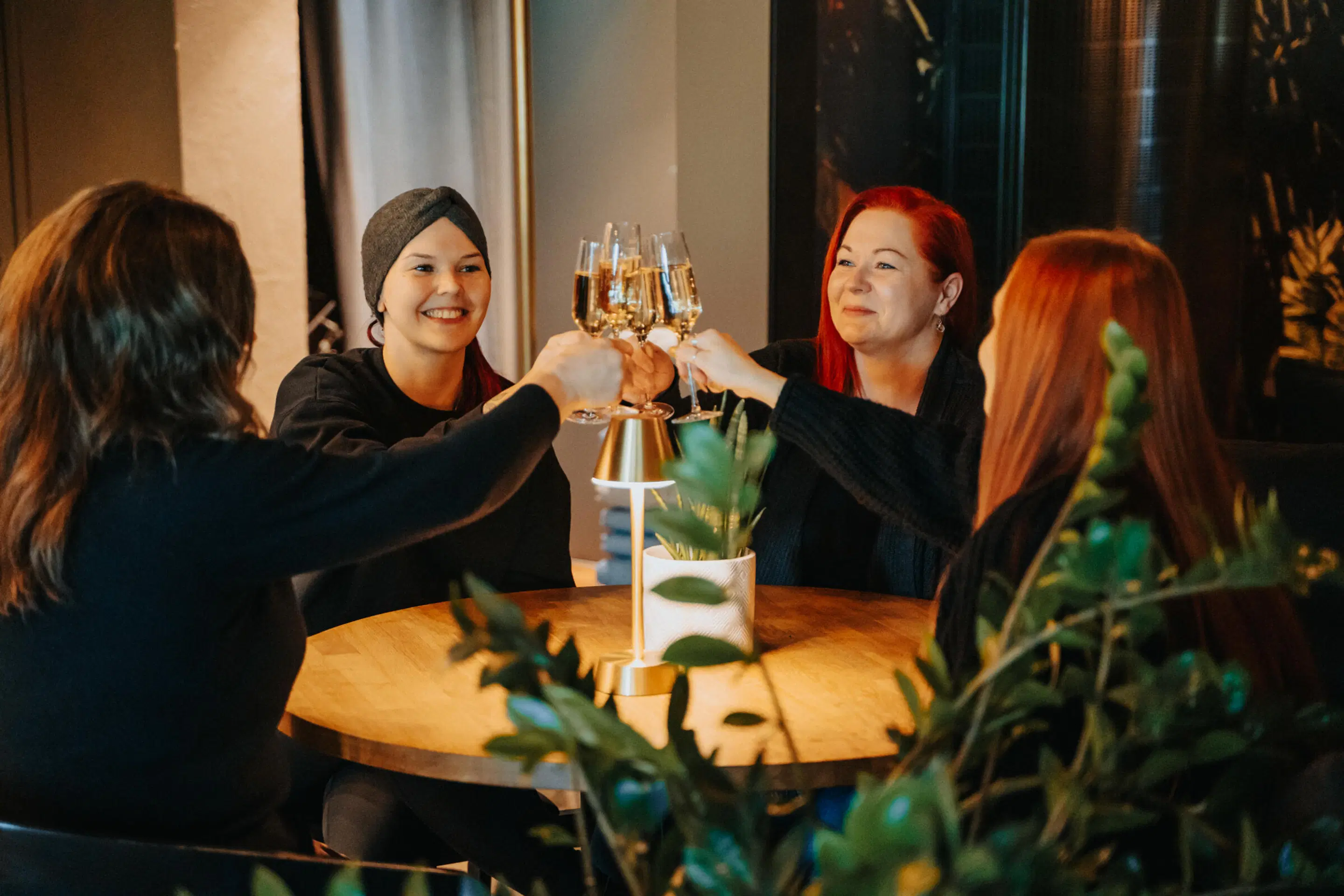 Four people toasting with champagne glasses around a round wooden table. A small table lamp and plant sit on the table.