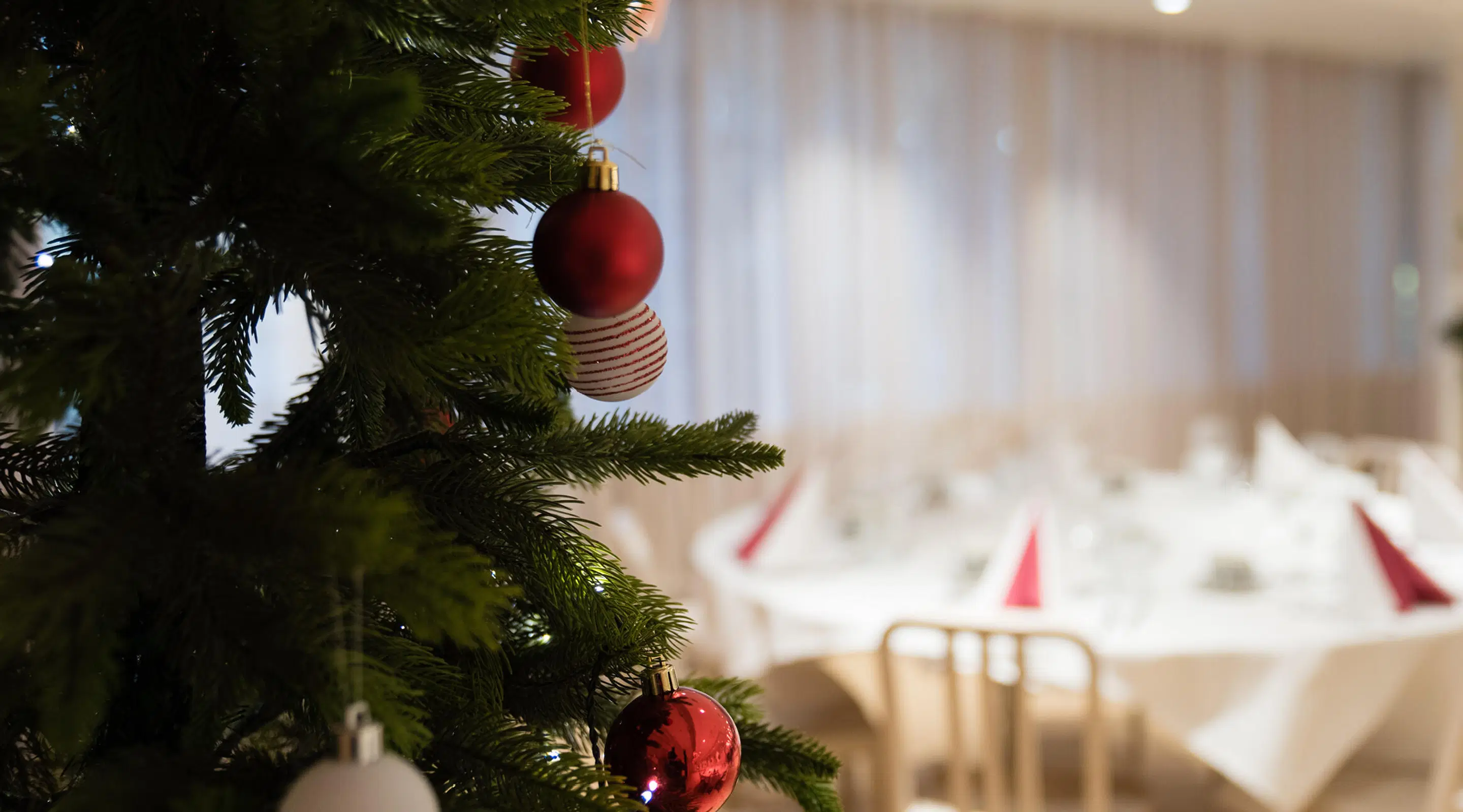Christmas tree with red and white ornaments in foreground, festive dining tables with red napkins in background.
