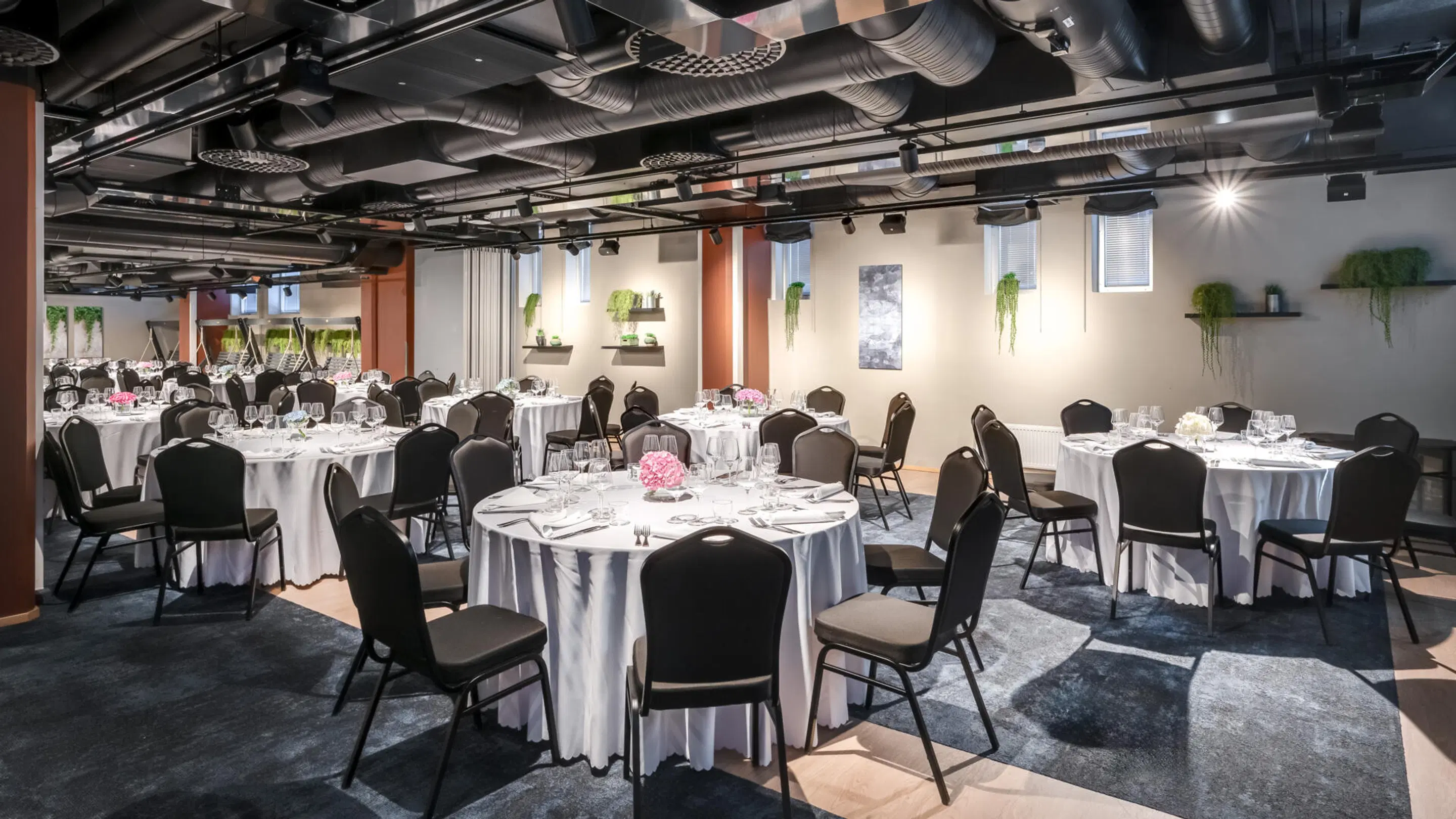 Event space with round tables, white tablecloths and black chairs. Green plants on walls, level floor and open pathways between tables.