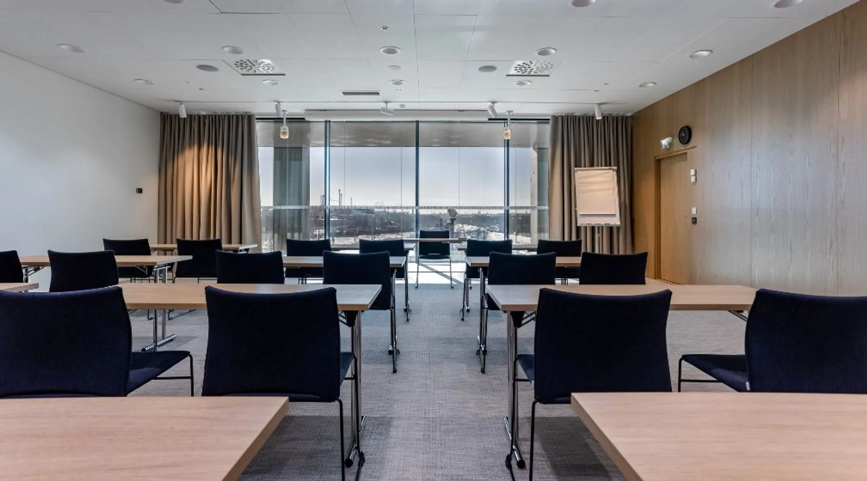 Conference room with rows of tables and chairs, large windows, curtains and flat carpet.