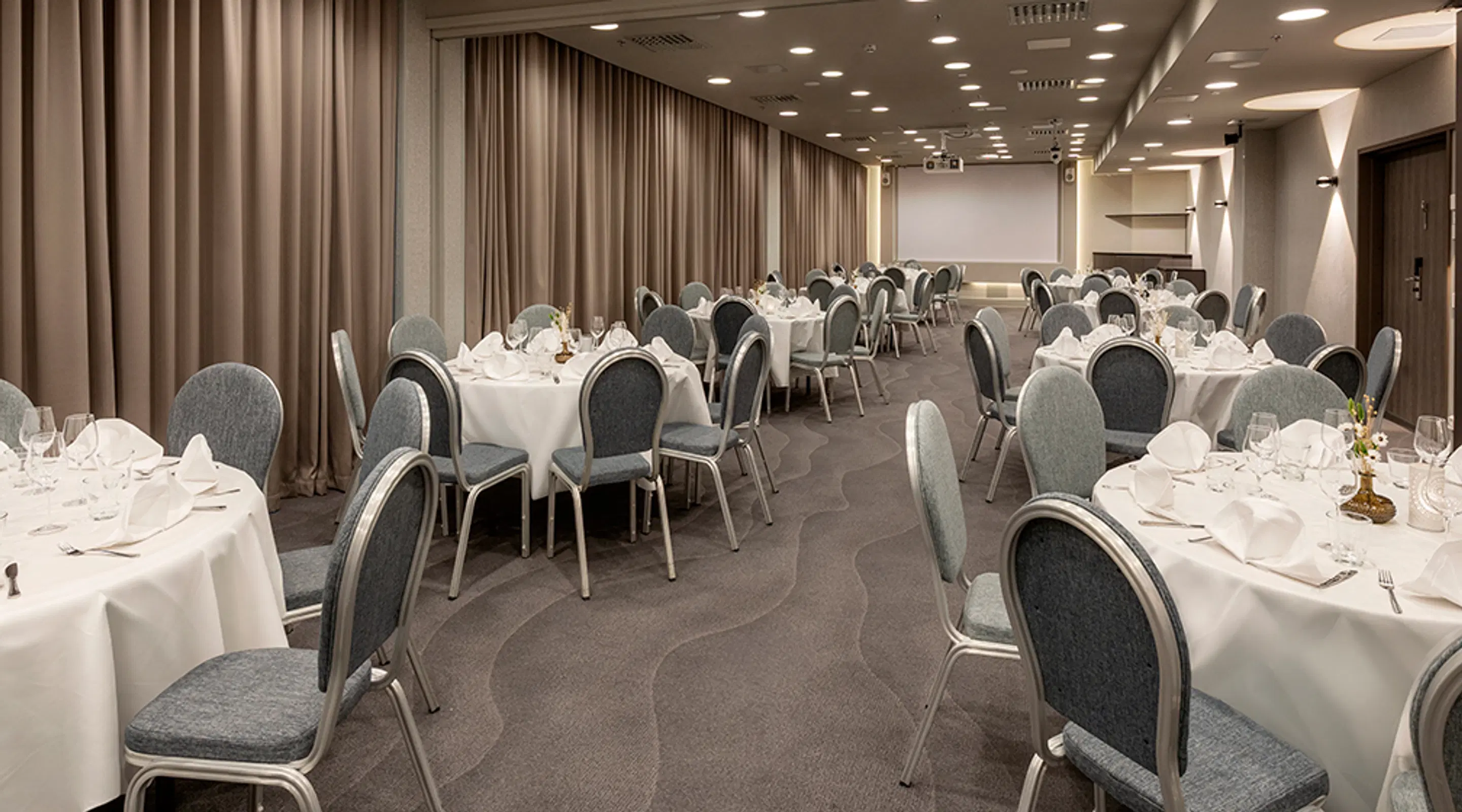 Banquet room with round tables in white linens and grey upholstered chairs. The floor has patterned carpet. A wide aisle runs through the middle. Ceiling spotlights and curtains.
