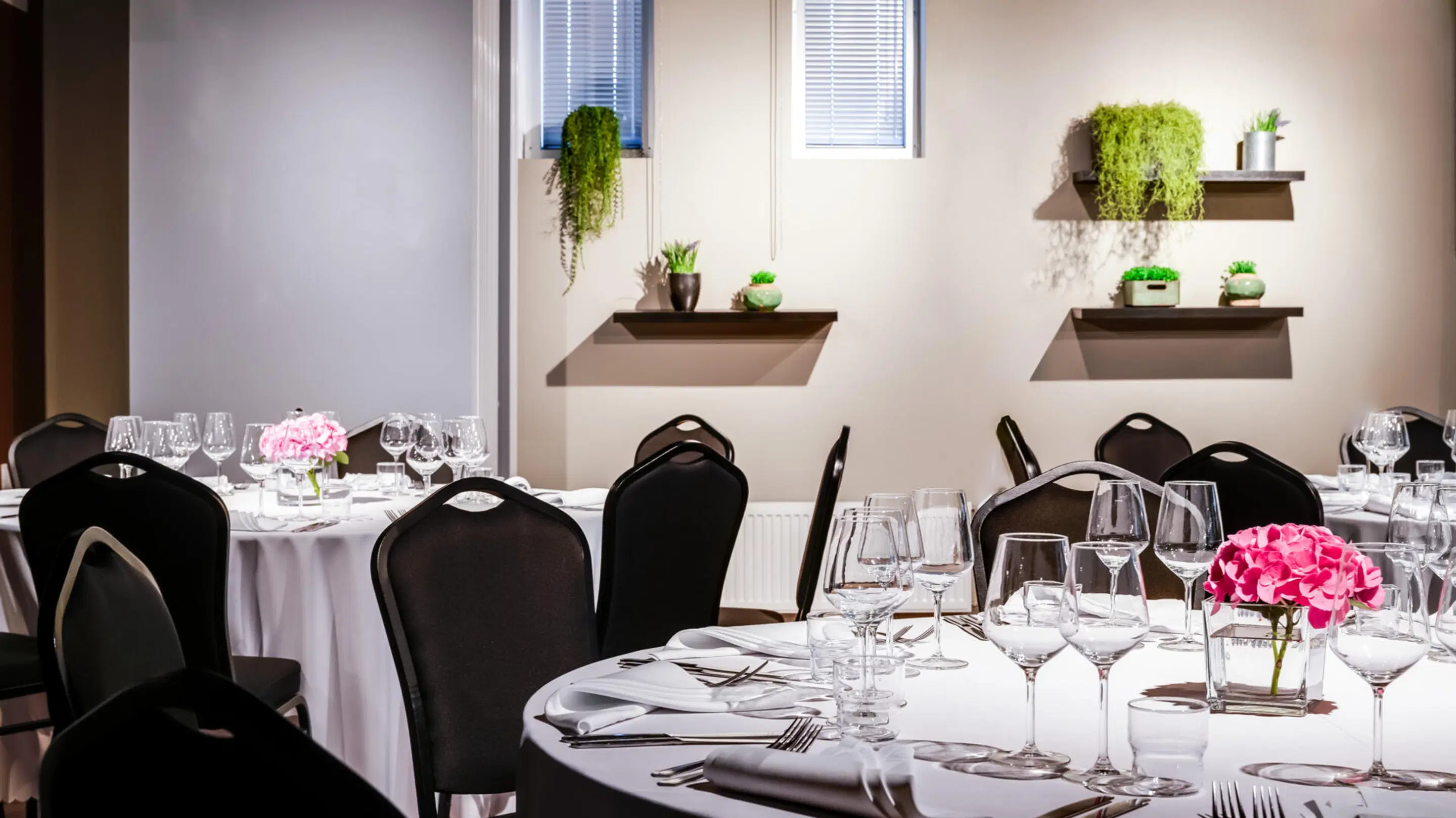 Banquet room with round tables covered in white tablecloths, black chairs and wine glasses. Green plants on wall shelves, pink flowers in vases on tables.
