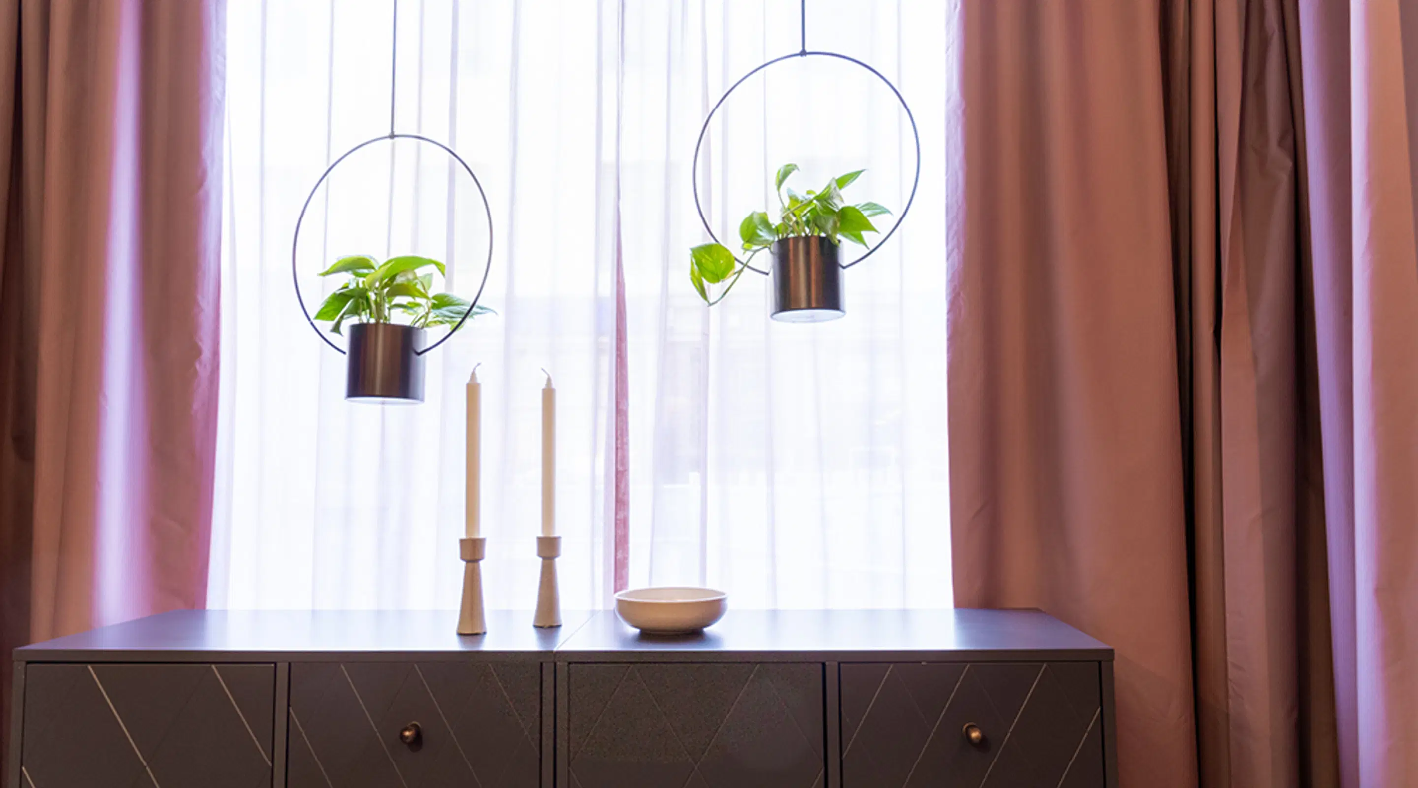 Dresser in front of window with two white candles, wooden bowl, and two hanging houseplants in metal planters.