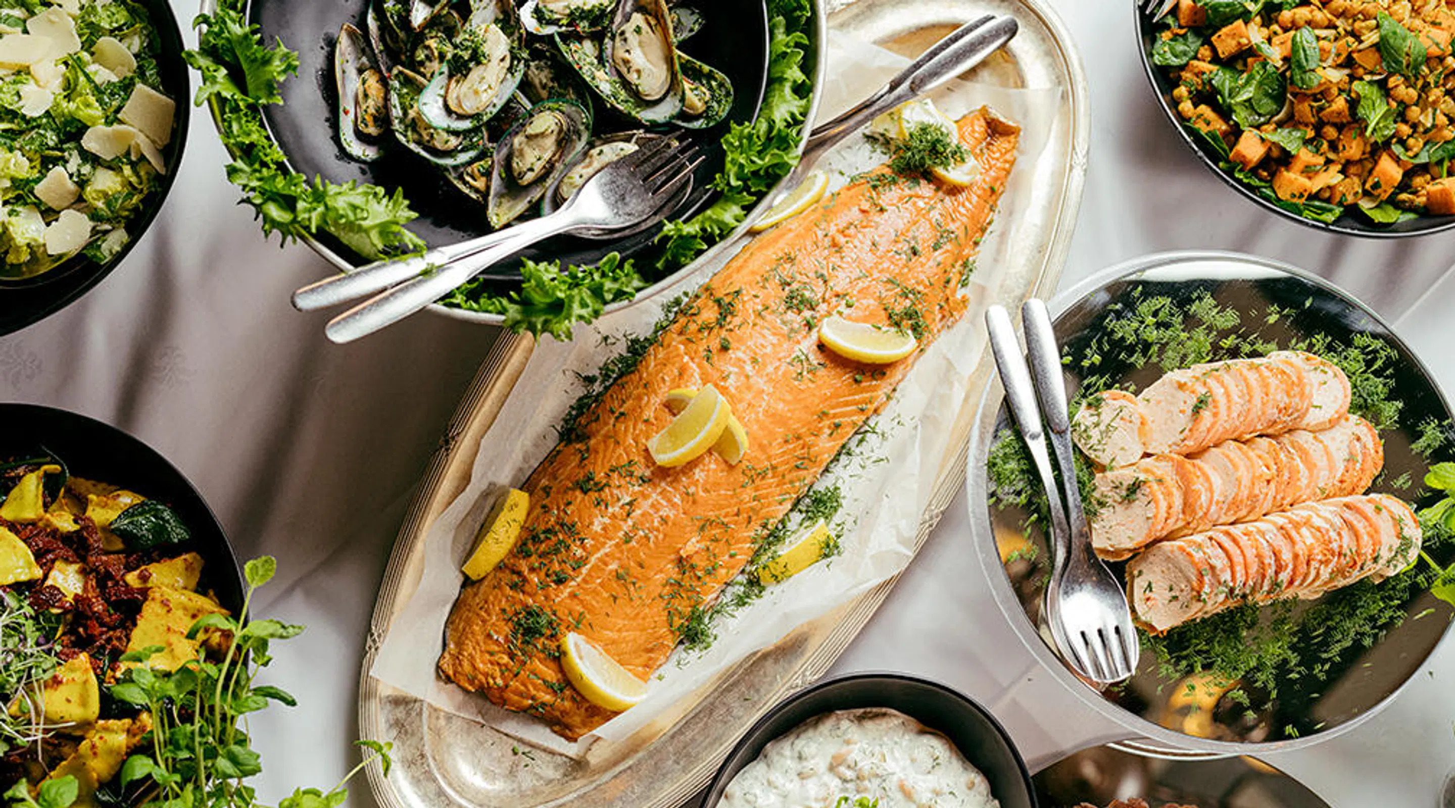Buffébord med vit duk med olika maträtter, en hel stekt lax på vitt fat i mitten.