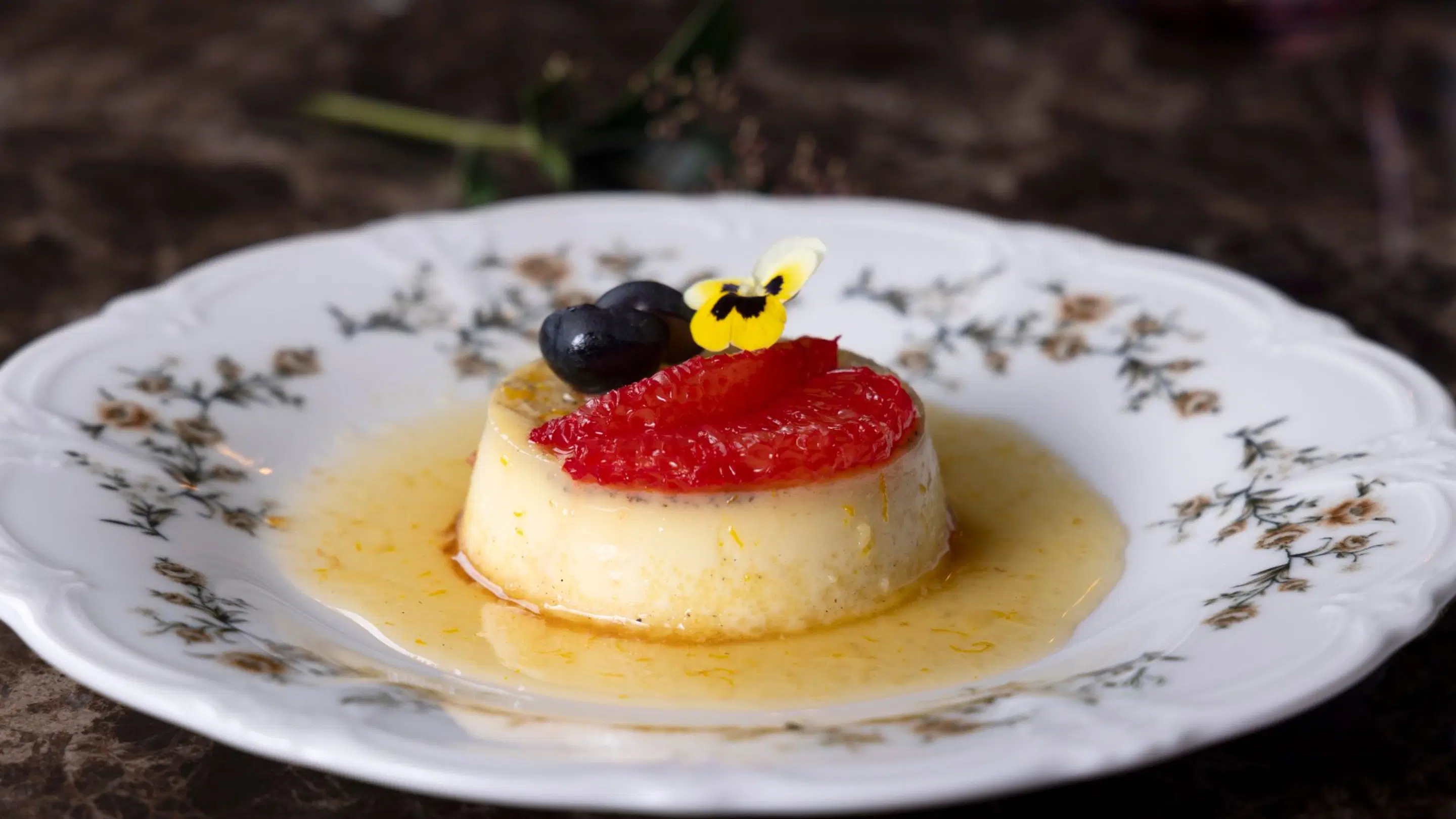 Panna cotta dessert on a decorative porcelain plate, topped with strawberry, blueberry and a yellow edible flower in a light sauce.