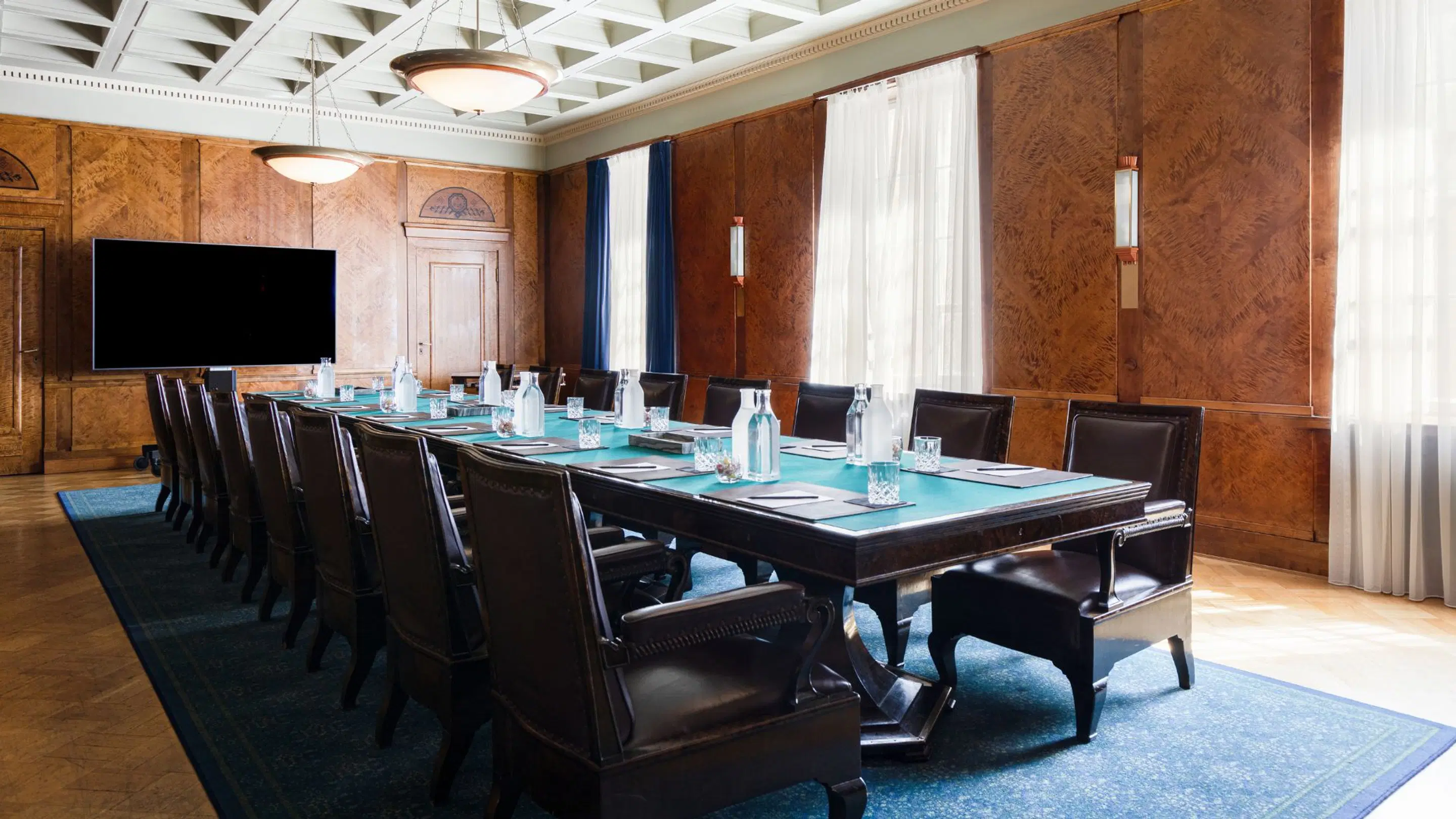 Elegant conference room with wood-paneled walls. Long table with blue tabletop and dark leather armchairs. Large windows, blue carpet and flat screen on wall.