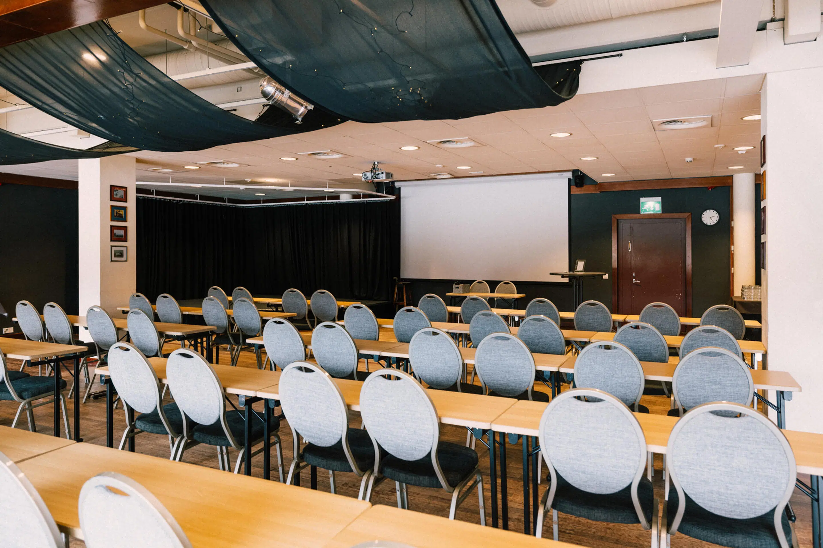 Konferensrum med bordsrader och stolar, projektorduk framme och svart gardin på sidan. Jämn belysning och brunt golv.